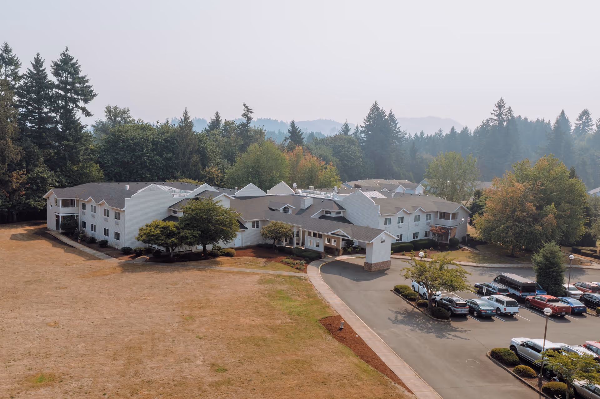 Aerial view of Wiley Creek Senior Living facility showing a large, two-story white building surrounded by trees and a parking lot with several cars. The building is set against a backdrop of forested hills under a hazy sky.