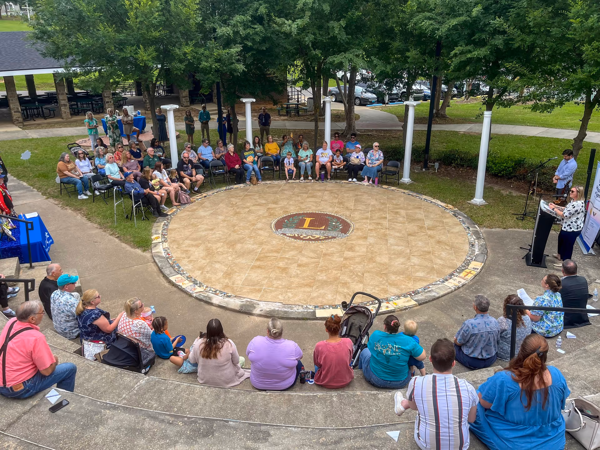 Outdoor circular courtyard with people seated around a tiled center, listening to a speaker.