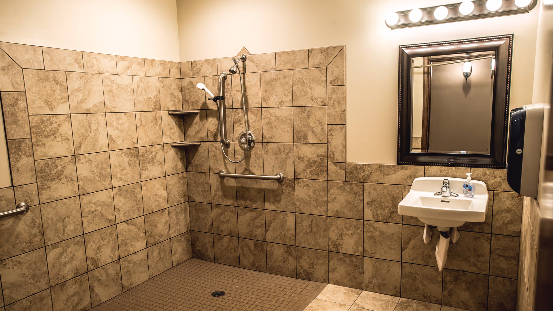 Tile-lined accessible bathroom with a roll-in shower, grab bars, a wall-mounted sink and mirror.