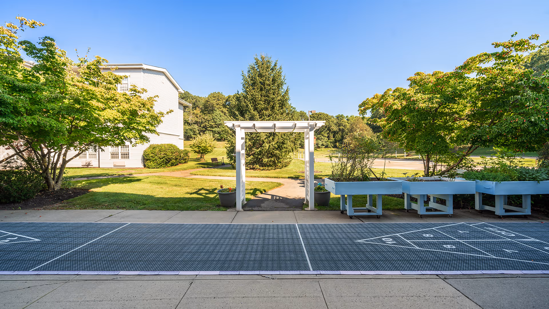 Outdoor area at Atria Hertlin Place featuring a white pergola entrance, green trees, shrubs, and raised garden beds on the right. There is a shuffleboard court in the foreground and a white building partially visible on the left under a clear blue sky.
