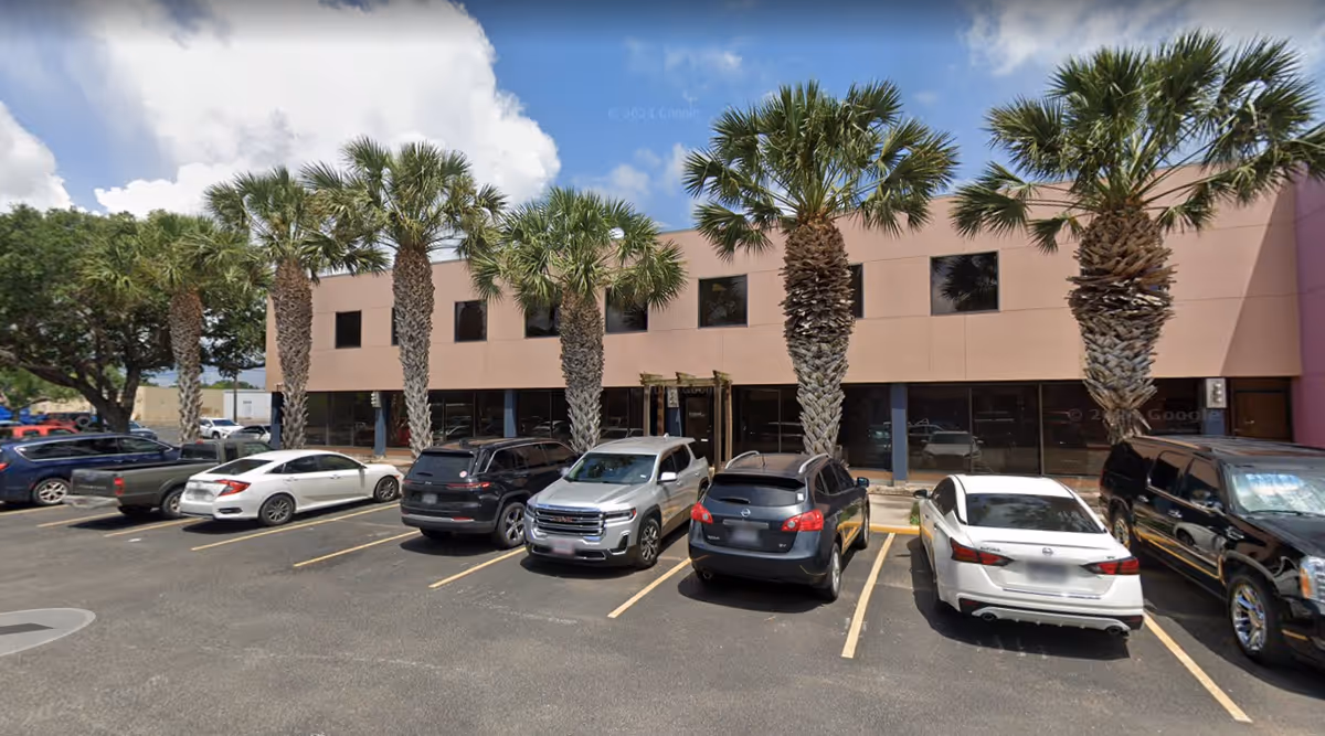 Exterior view of a beige two-story building with tall palm trees and parked cars.