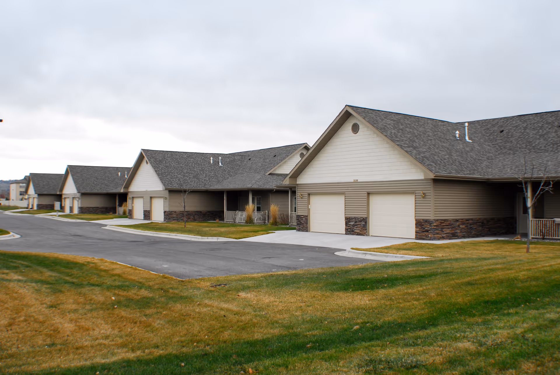 Row of beige retirement community buildings with garages and green lawns along a curved street.