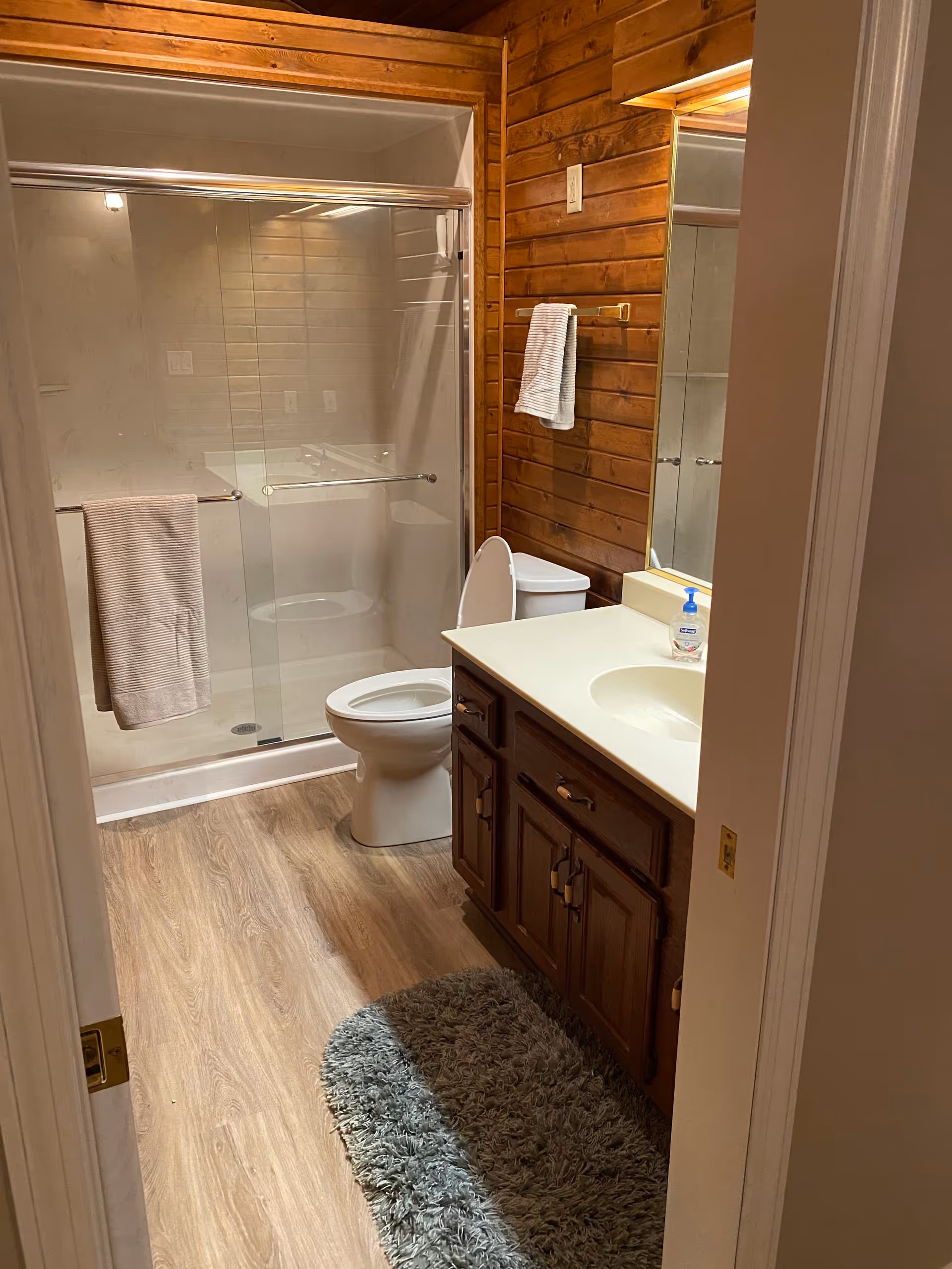 Wood-paneled bathroom with glass shower, vanity, toilet, and rug.