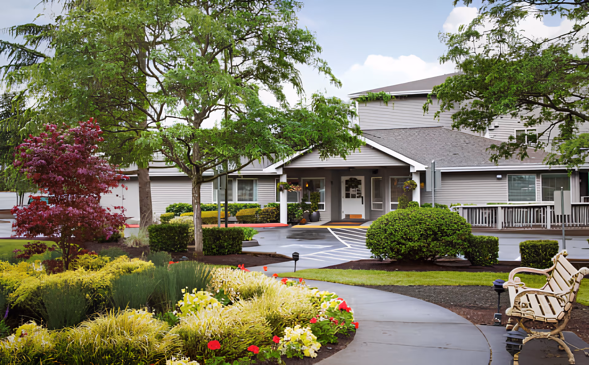 Entrance of a memory care facility with a covered front porch, walkway, benches, and landscaped gardens.