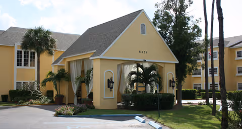 Exterior view of a yellow senior living facility building with a covered entrance, palm trees, and a driveway with parking spaces in front.