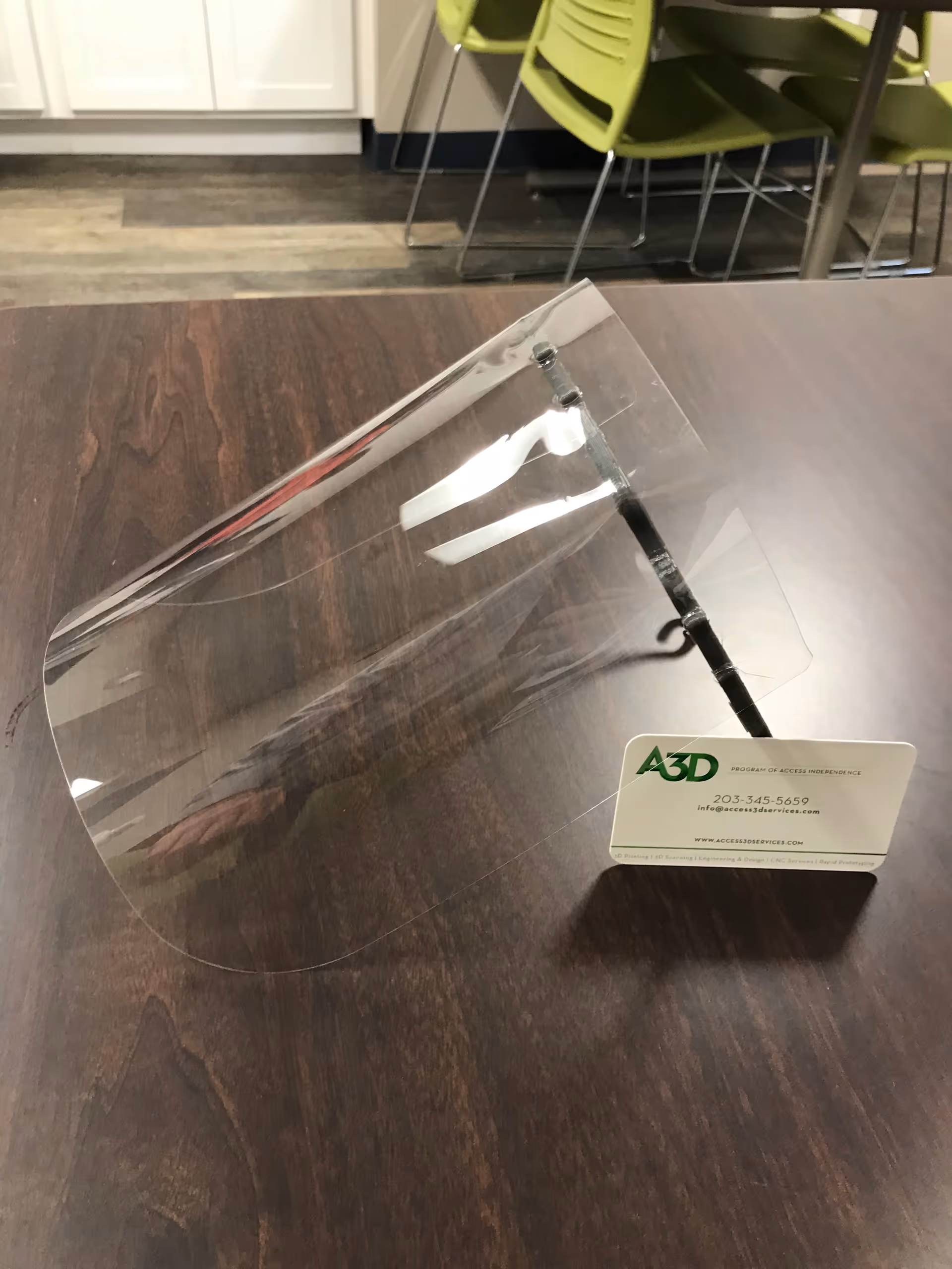 Clear face shield on a dark wooden table with green chairs in background