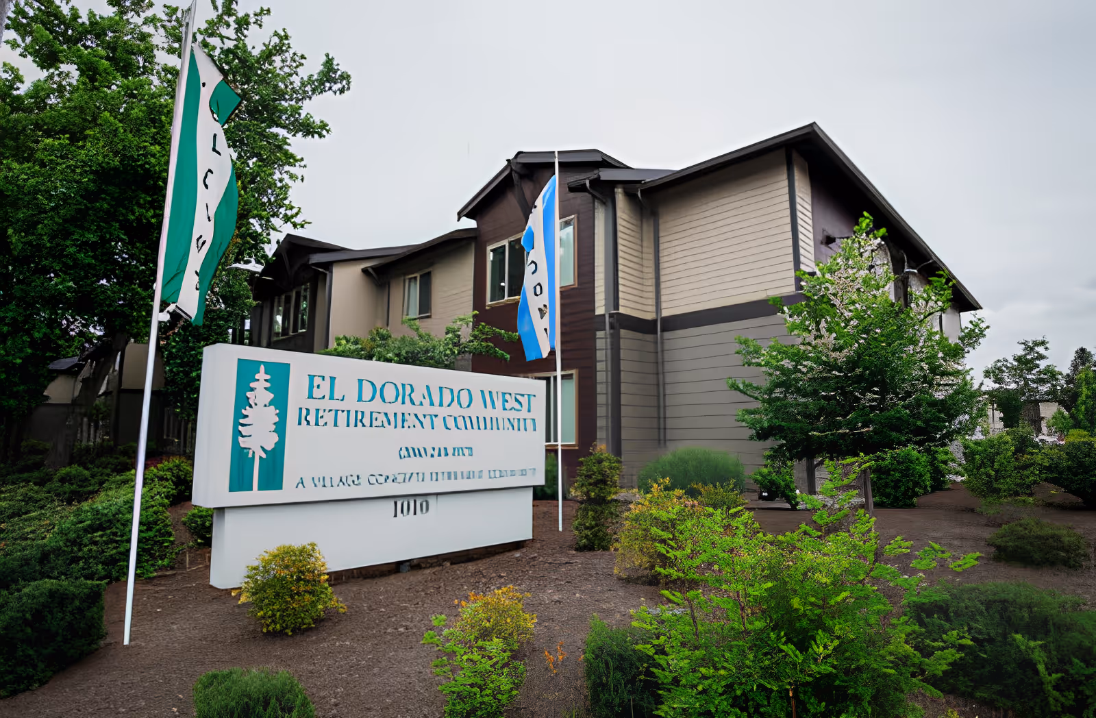 Exterior view of the El Dorado West Retirement Community building with a large sign in front displaying the community name. The building is surrounded by green shrubs and trees under a cloudy sky.