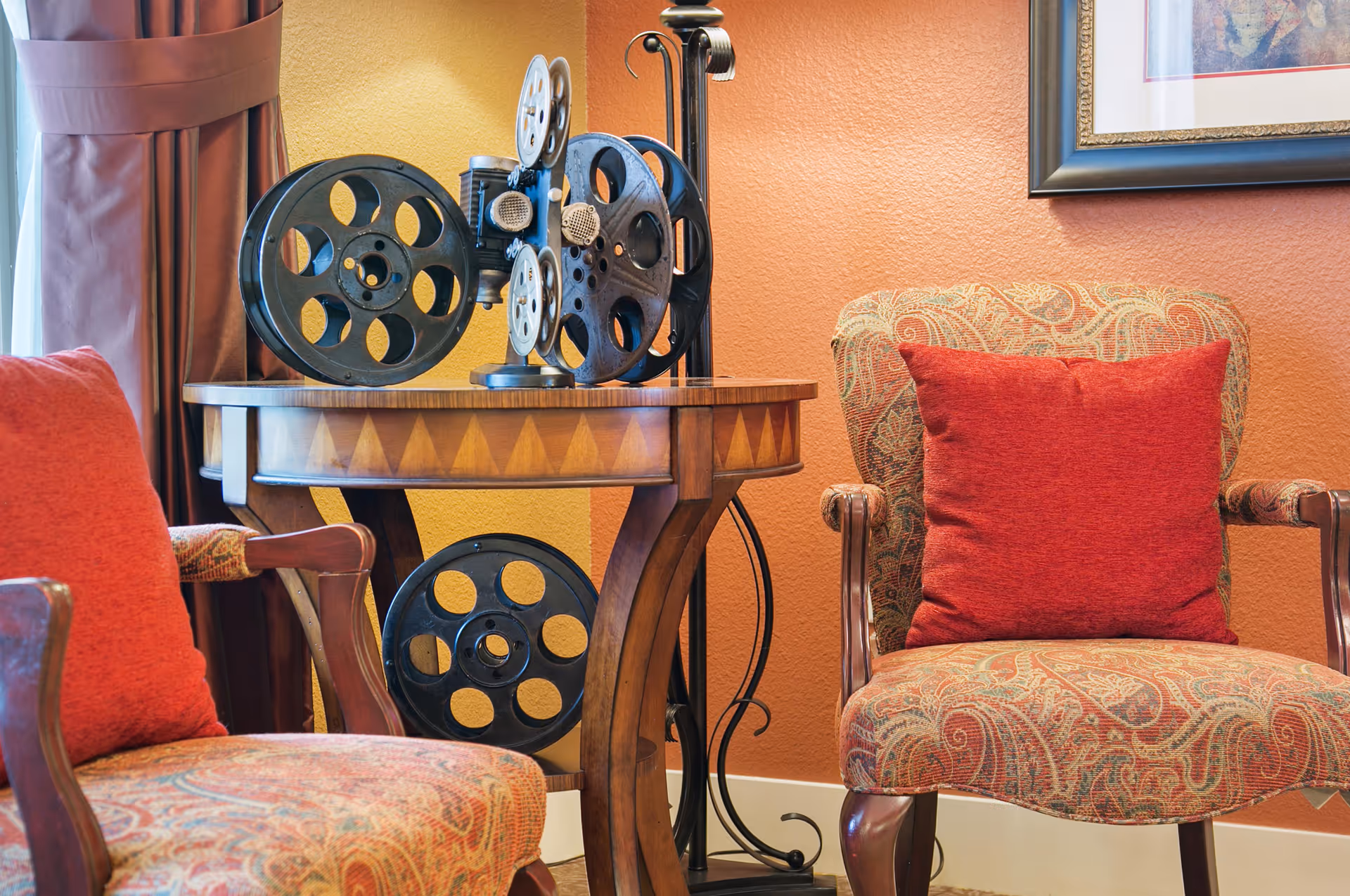 A cozy sitting area with two patterned armchairs, each with a red cushion. Between the chairs is a wooden side table holding vintage film reels. The background features an orange textured wall with a framed picture partially visible.