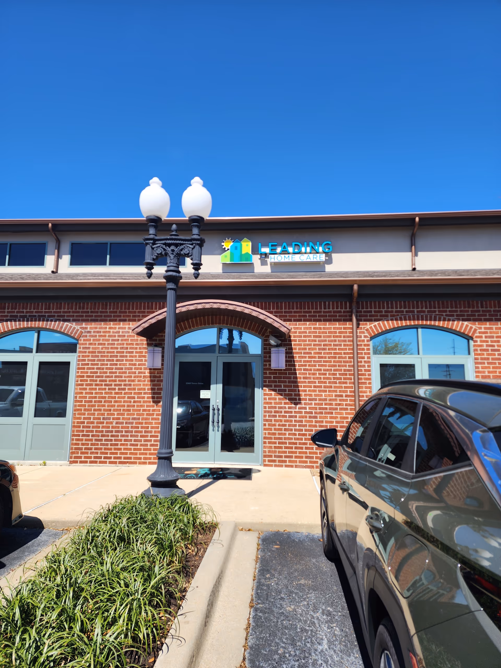 Brick facility exterior with front entrance, sign reading Leading Home Care Monroe, and a parked car.
