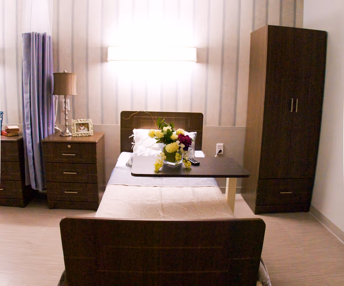 A neatly arranged bedroom in a senior living facility featuring a single bed with white bedding and a floral arrangement on an overbed table. The room includes a bedside table with a lamp and a framed photo, a tall wardrobe, and a window with purple curtains. The walls have a subtle striped pattern and a bright wall light above the bed.