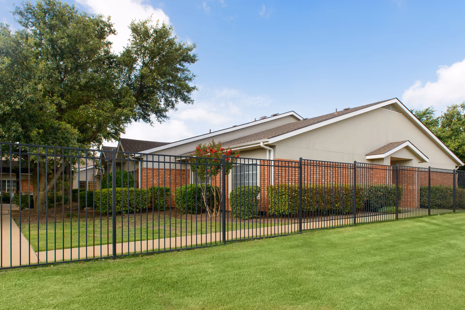 Exterior view of a single-story building with brick and beige siding, surrounded by a black metal fence and green lawn, with trees and shrubs near the building under a blue sky with some clouds.