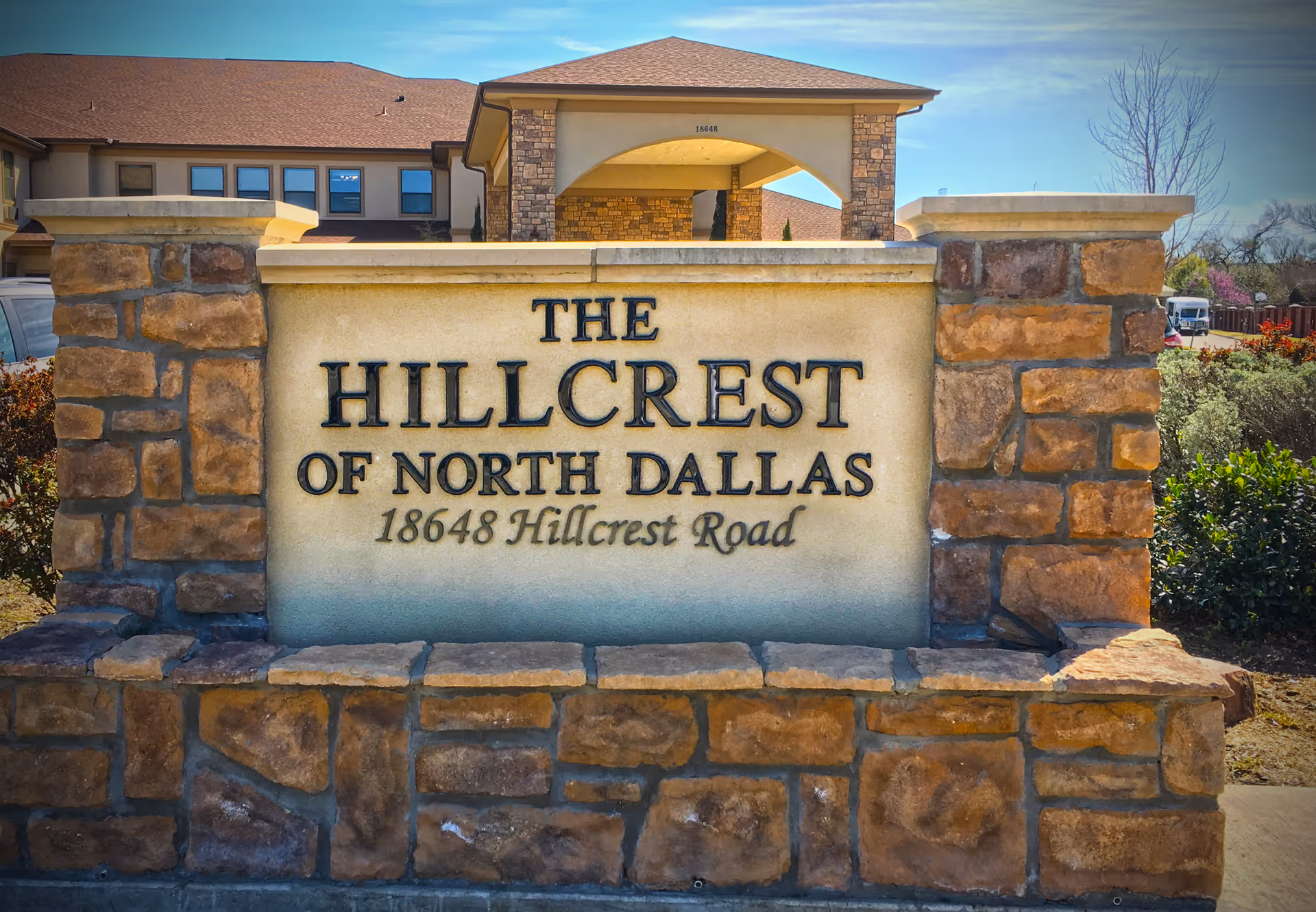 Stone sign at the entrance of The Hillcrest of North Dallas facility with the address 18648 Hillcrest Road, with part of the building and landscaping visible in the background under a clear blue sky.