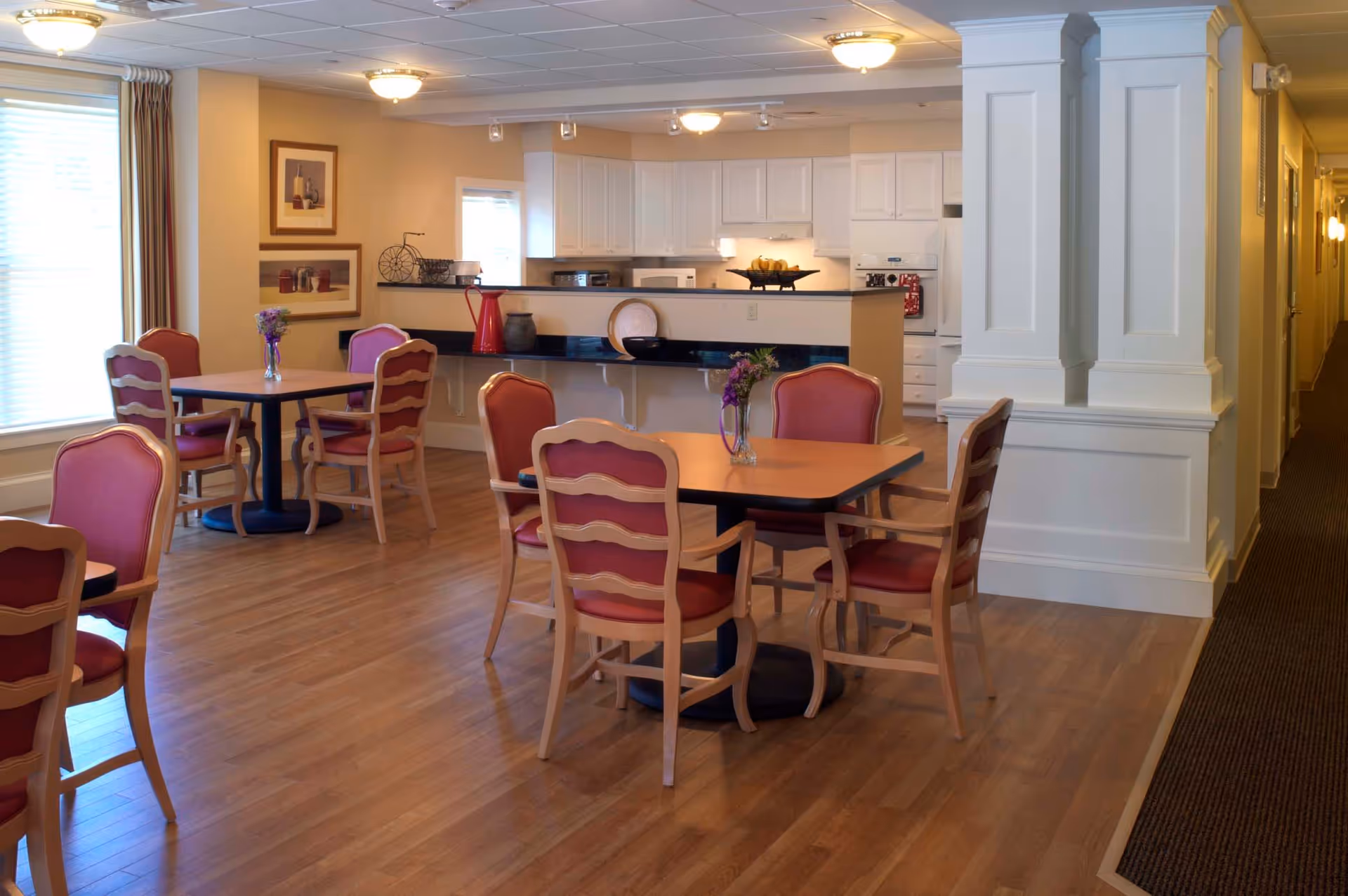 A dining area in a senior living facility with wooden floors, several tables surrounded by wooden chairs with red cushions, and a small vase with flowers on each table. In the background, there is a kitchen area with white cabinets, a countertop, and various kitchen items. The room is well-lit with ceiling lights and natural light from a window with blinds and curtains.