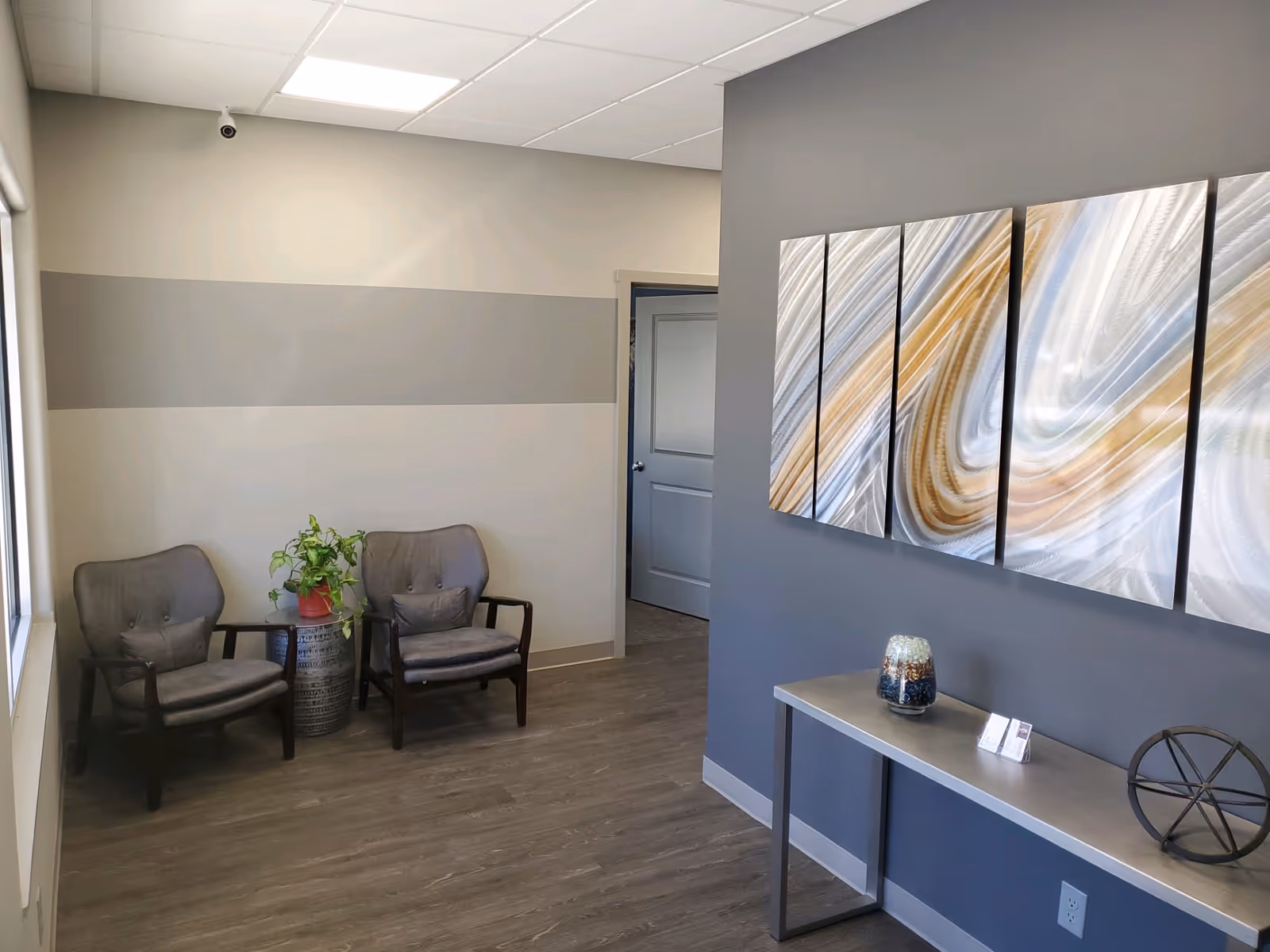 Cozy waiting area with two gray chairs, potted plant, and abstract triptych art.