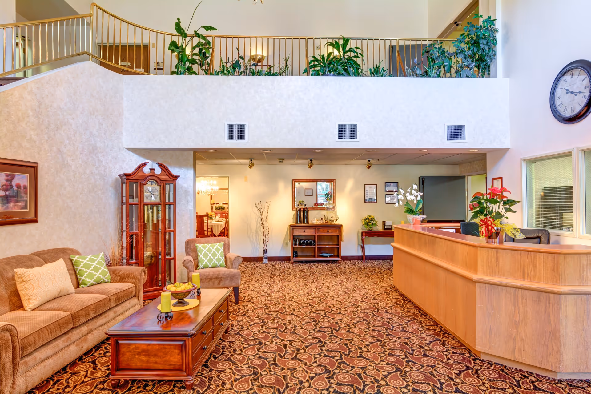 A bright and welcoming lobby area with a brown sofa and armchair featuring green and beige cushions, a wooden coffee table with decorative items, a tall grandfather clock, and a wooden reception desk with plants. The space has patterned carpet flooring, a large wall clock, and an upper level with plants along the railing.
