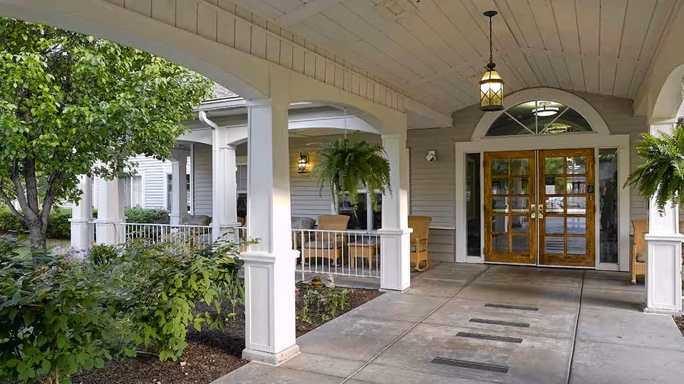 Covered entrance area of a senior living facility with white pillars, hanging plants, wicker chairs, and a wooden double door with glass panels. There are green bushes and a tree on the left side under the covered porch.