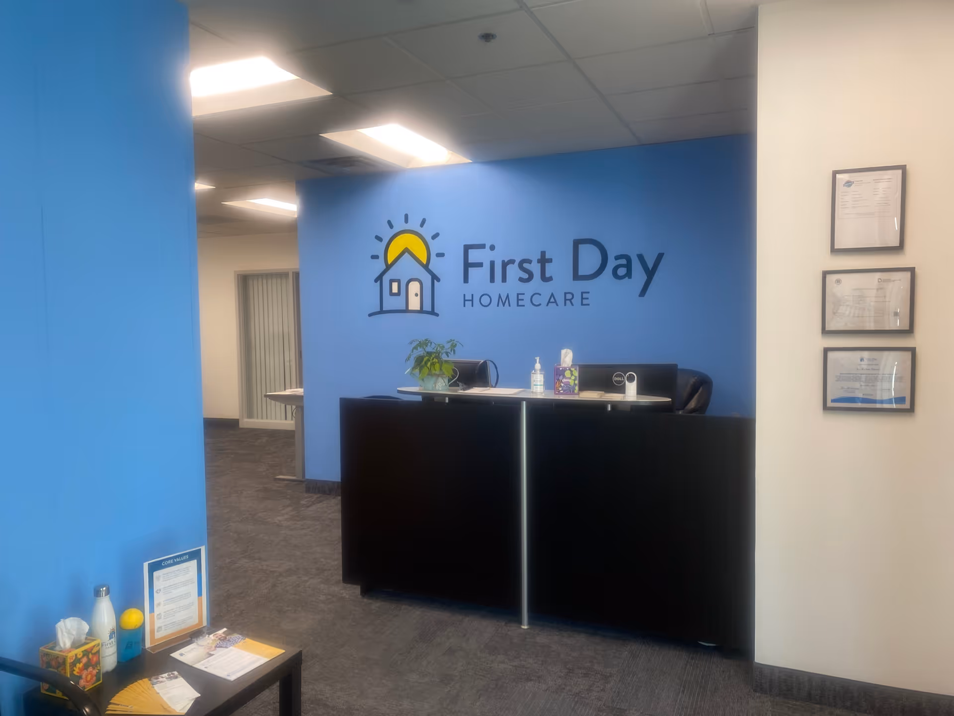 Reception desk in a blue-walled lobby featuring First Day Homecare logo and framed certificates.