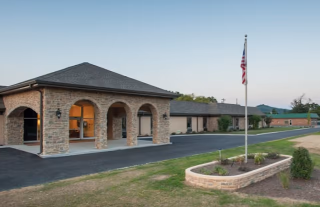 Single-story brick senior living facility entrance with arched portico, driveway, landscaped planter and an American flagpole.