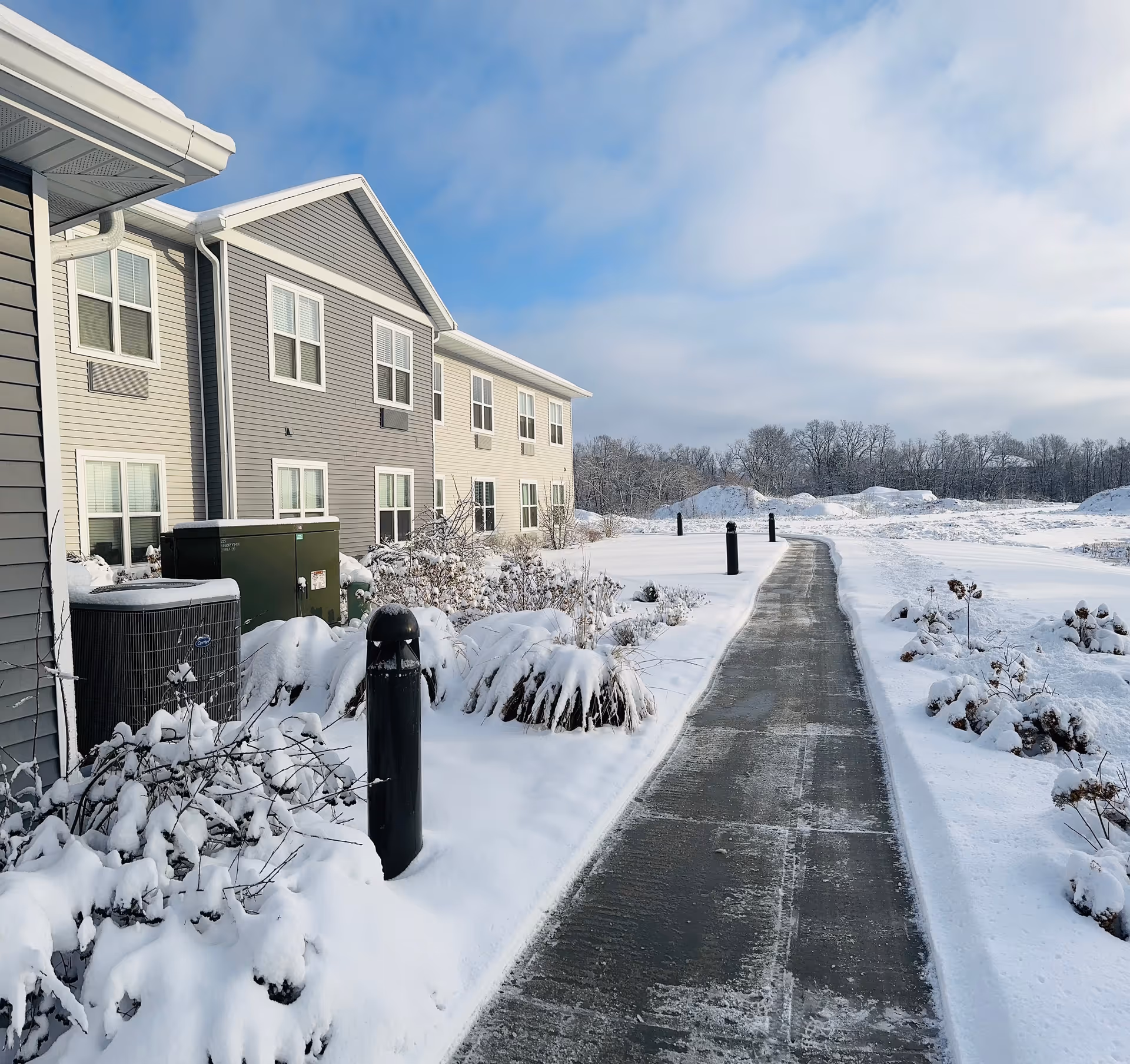 Snow-covered exterior of Riverwalk Senior Living with a cleared walkway beside buildings.