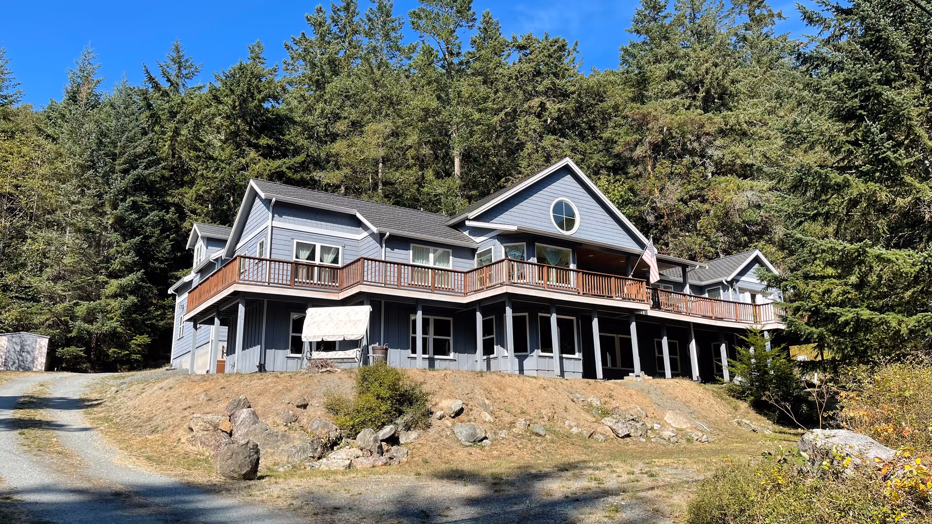 Blue-gray senior living facility with a wraparound deck beside tall trees.