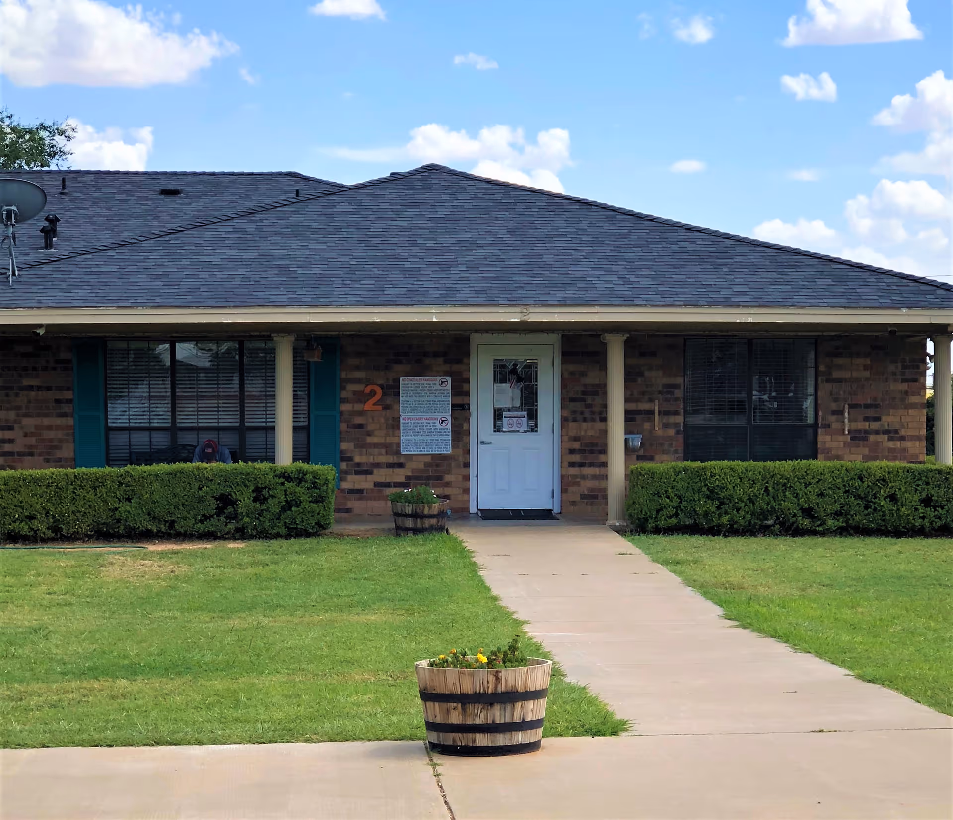 Front exterior view of a single-story brick building with a dark shingled roof, a white door in the center, two windows with green shutters on either side, trimmed bushes along the front, a concrete walkway leading to the door, and a wooden planter with flowers in the foreground under a partly cloudy sky.