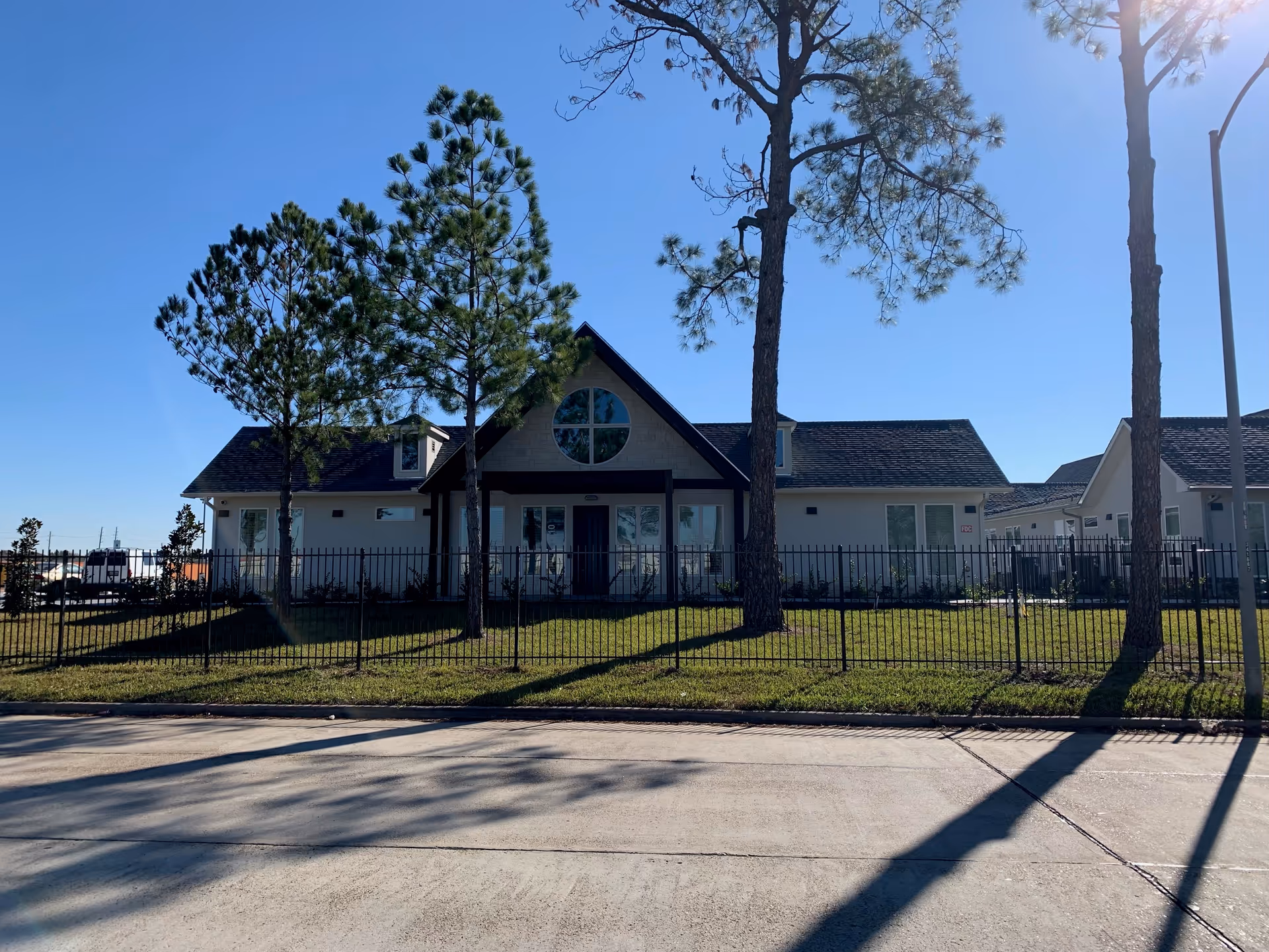 Front exterior view of a single-story assisted living facility building with a triangular roof and a large circular window above the entrance. The building is surrounded by a black metal fence and several tall pine trees, with a clear blue sky in the background.