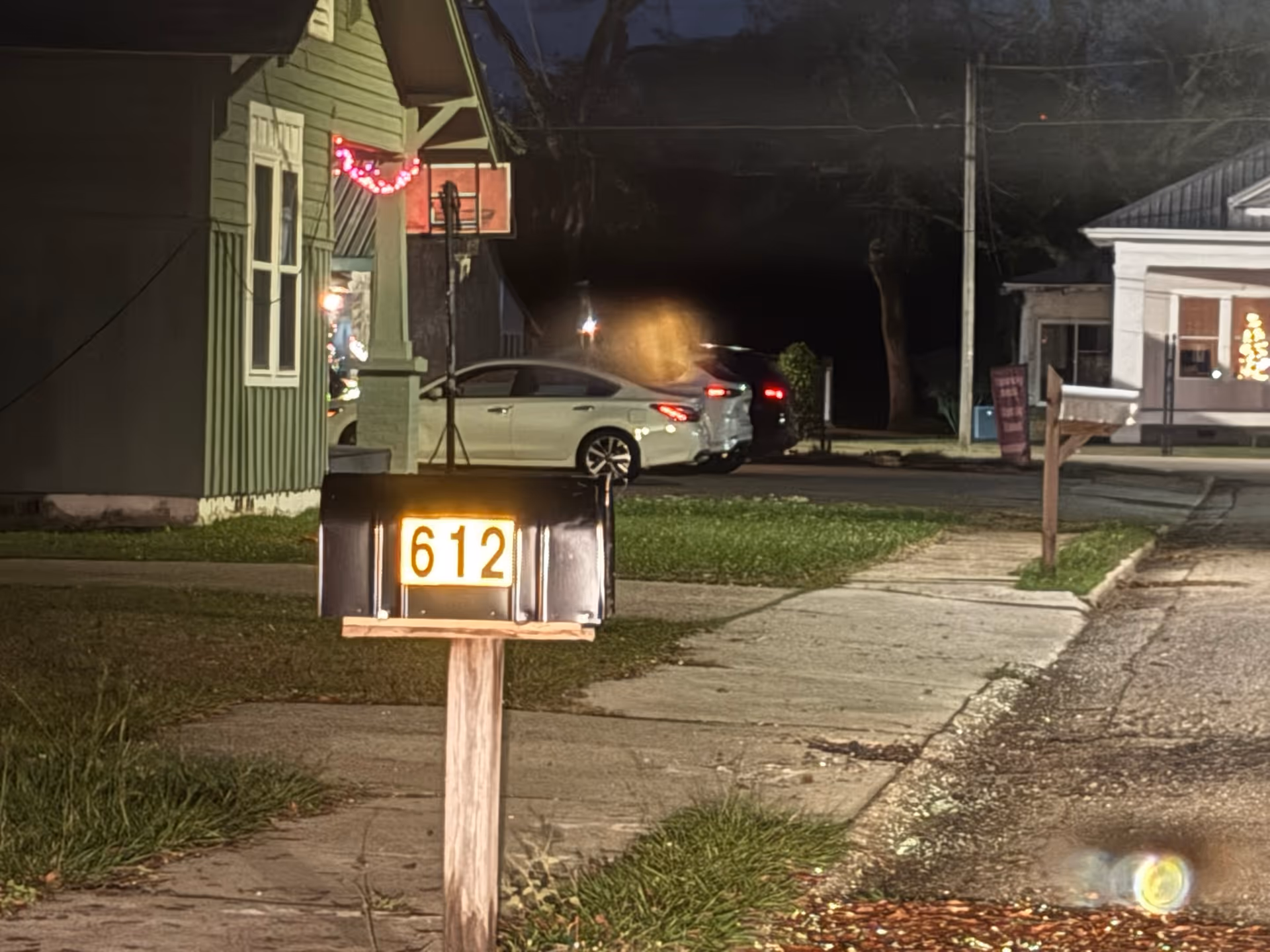 Night exterior view of a street with a mailbox, parked cars, and building fronts.