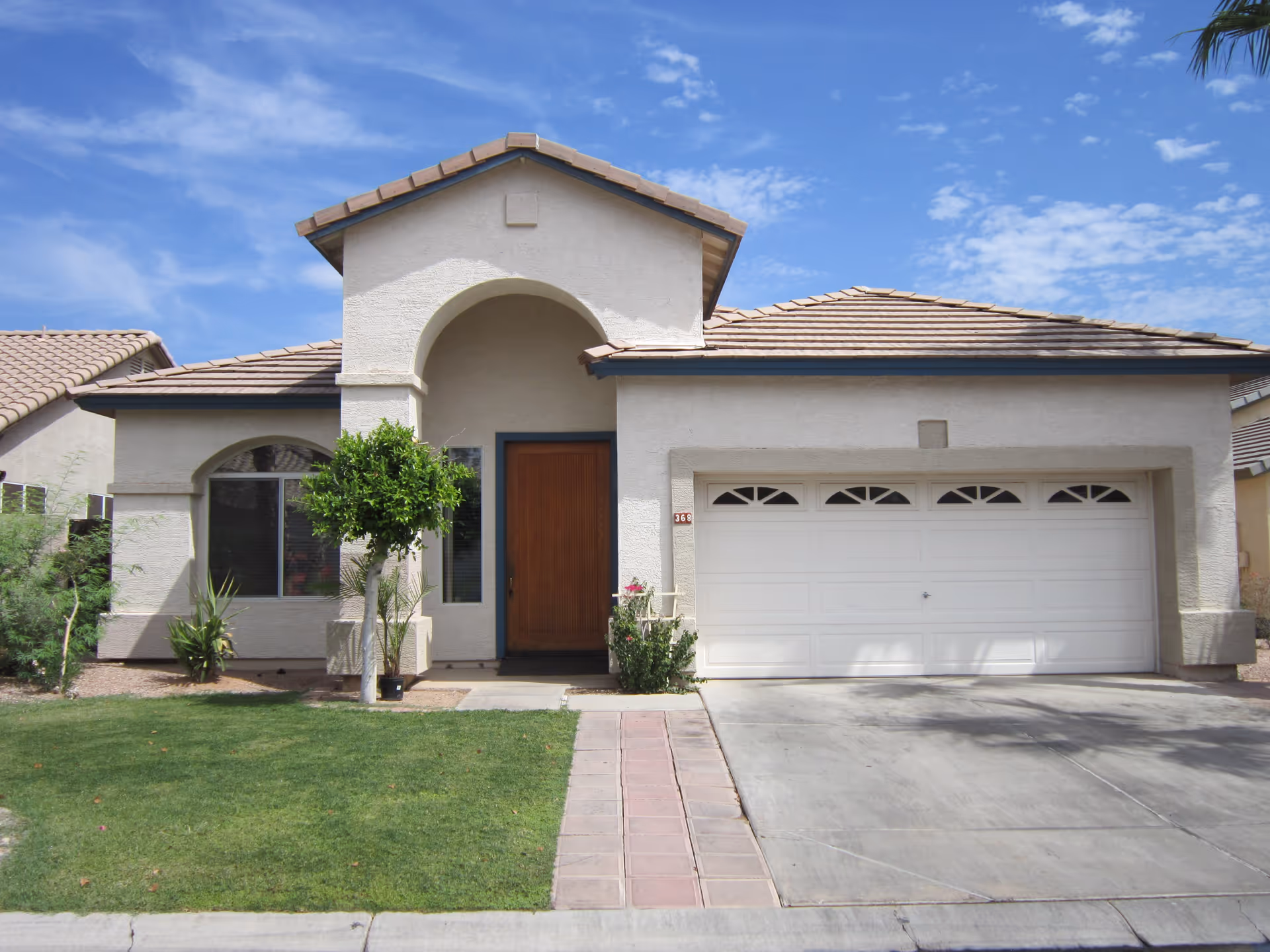 Front exterior of a single-story stucco house with an arched entryway, wooden door, two-car garage, lawn and driveway under a blue sky.