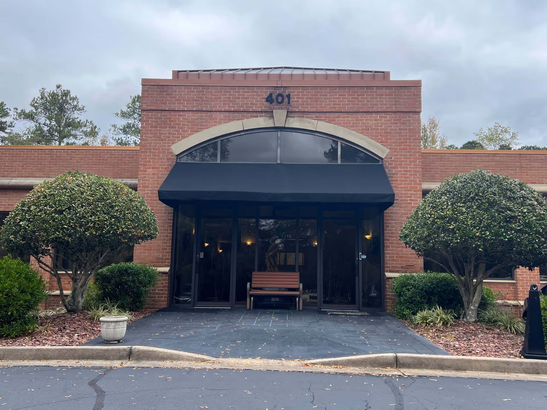 Brick building entrance with black awning, glass doors, bench, and manicured shrubs.