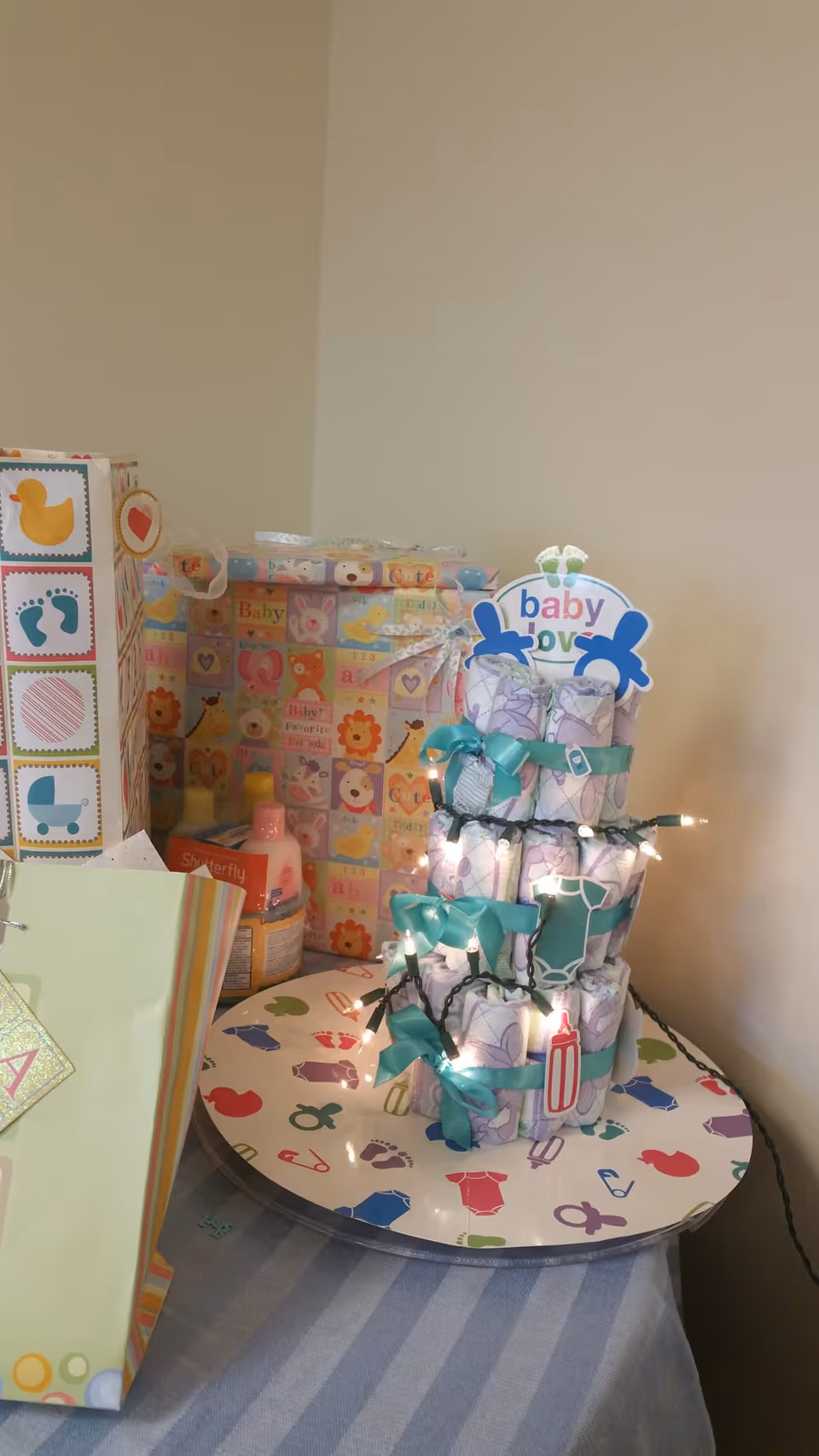 A table in a softly lit room displays a stacked diaper cake and baby-themed gifts.