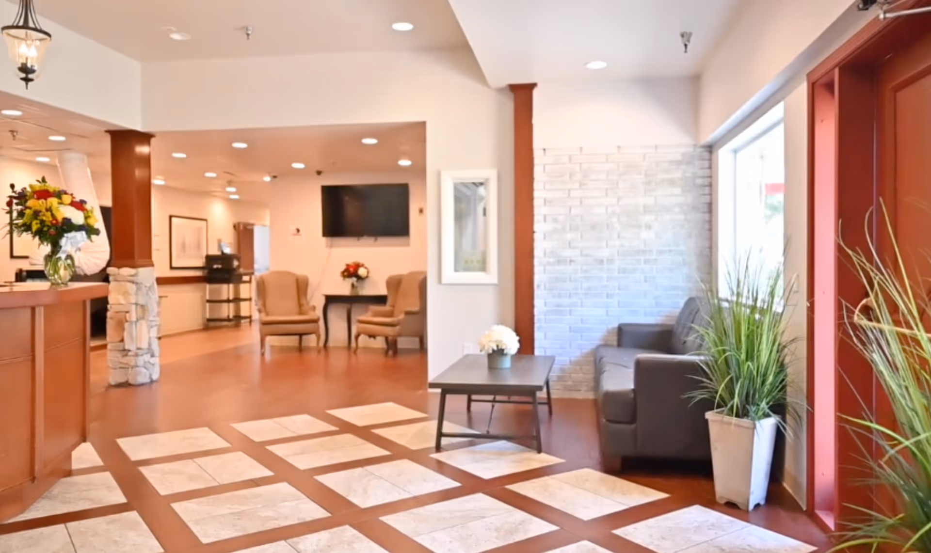A bright and welcoming healthcare center lobby with a wooden reception desk on the left adorned with a vase of flowers. The floor features a patterned tile design. There are two beige armchairs and a small table with flowers in the background, a wall-mounted TV, and a gray couch next to a window on the right. Potted plants are placed near the window and entrance door.