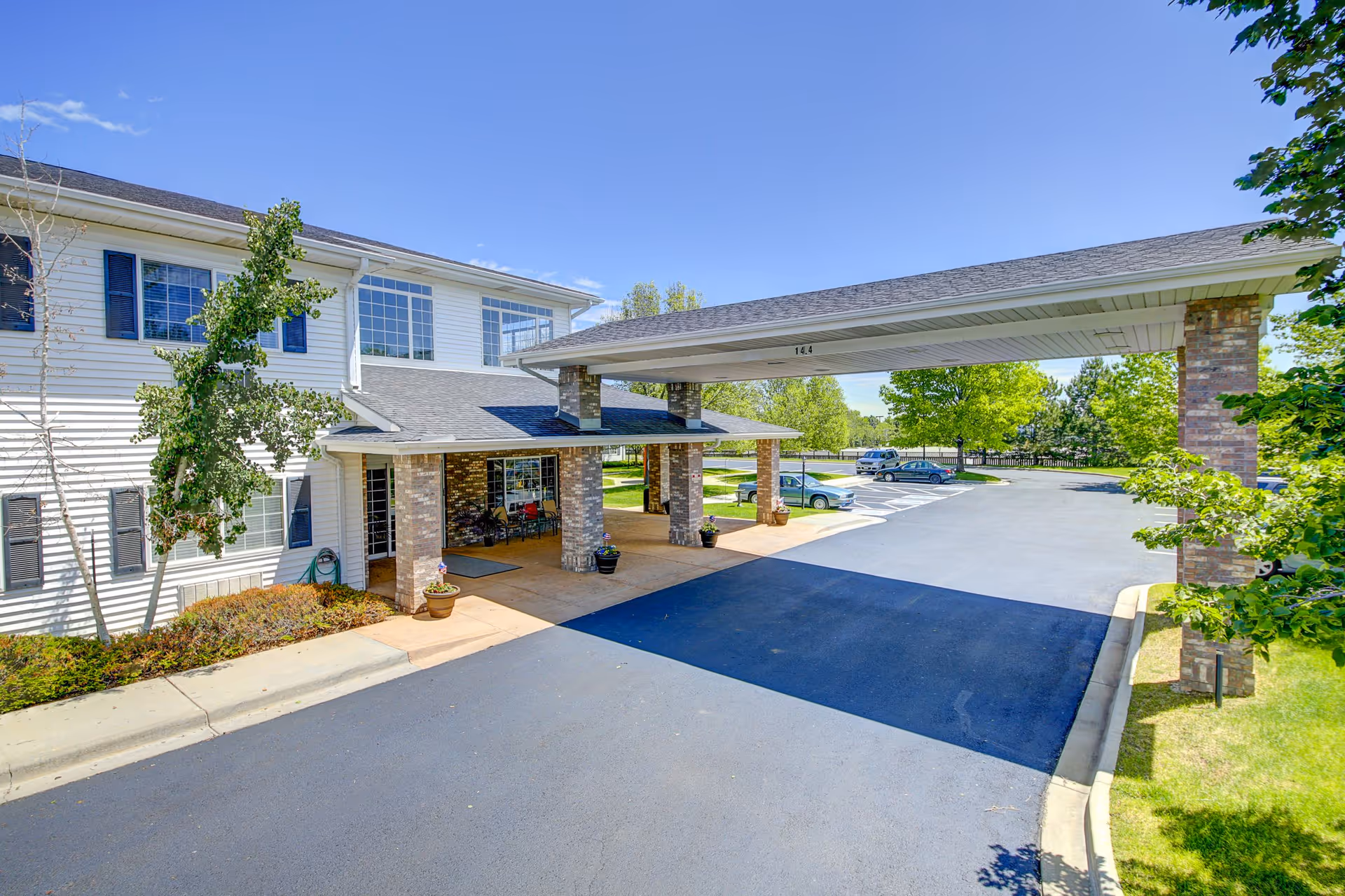 Exterior view of Waneka Park Assisted Living facility showing the entrance with a covered drop-off area supported by brick columns, a paved driveway, parked cars, and surrounding greenery under a clear blue sky.