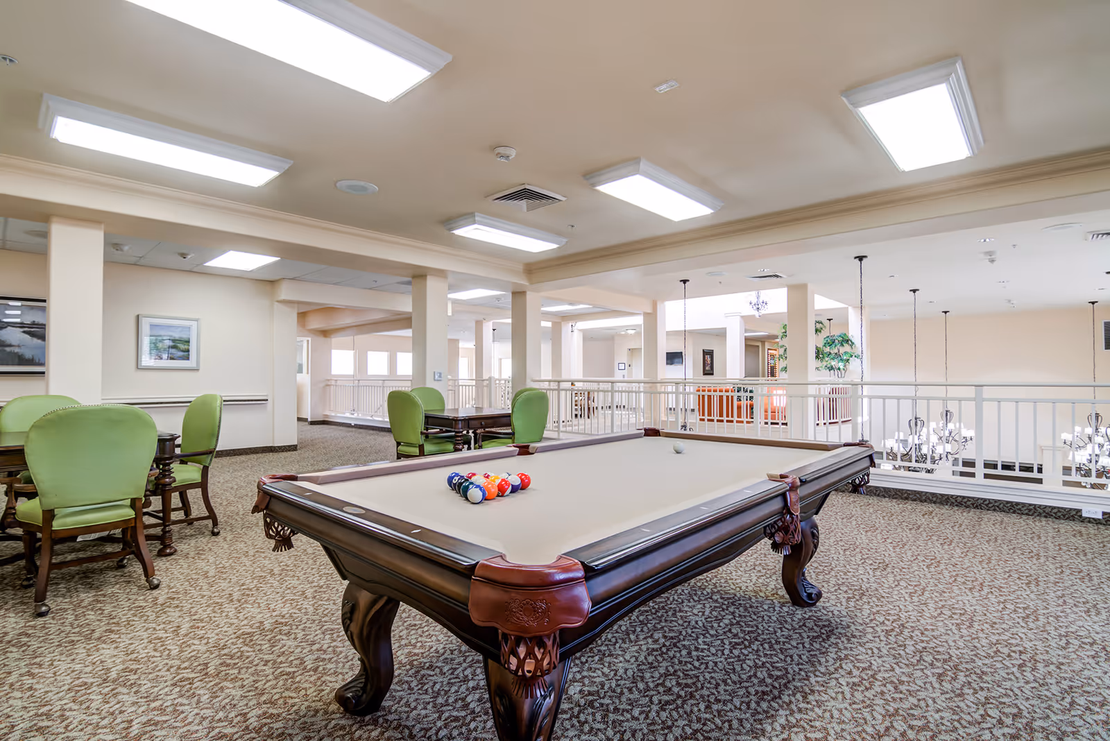 A spacious senior living facility common area featuring a billiards table with balls arranged for a game. Surrounding the table are several green upholstered chairs and wooden tables. The room is well-lit with ceiling lights and has beige walls and patterned carpet. A railing overlooks a lower level with chandeliers visible below.