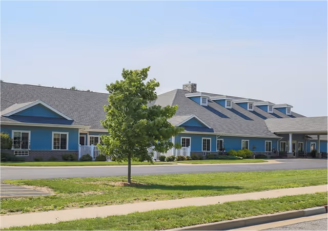 Blue single-story senior living building with sidewalk, lawn, and a tree in foreground.