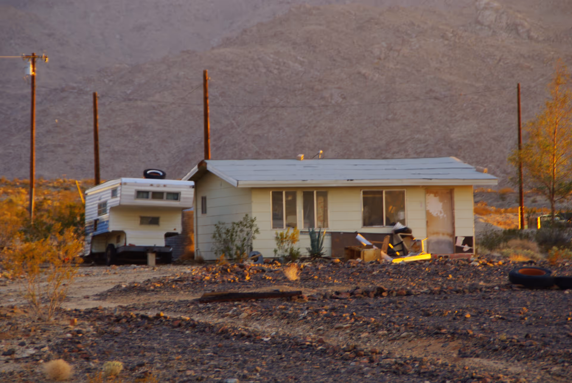 Desert landscape with a weathered trailer house and scattered debris.