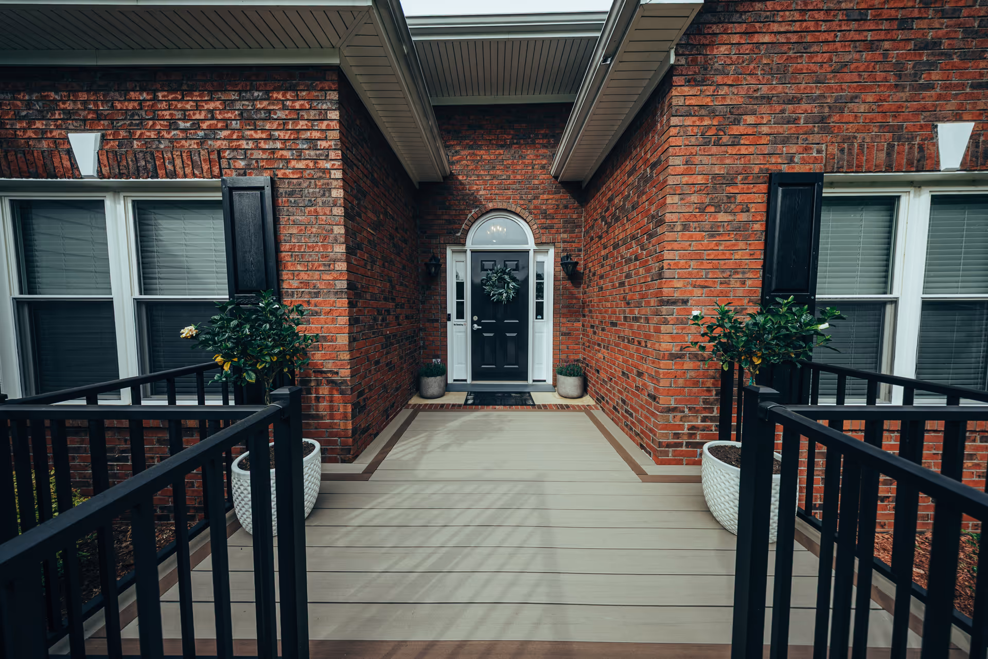 Brick building entrance with a black front door, wreath, and potted plants on a wooden porch.