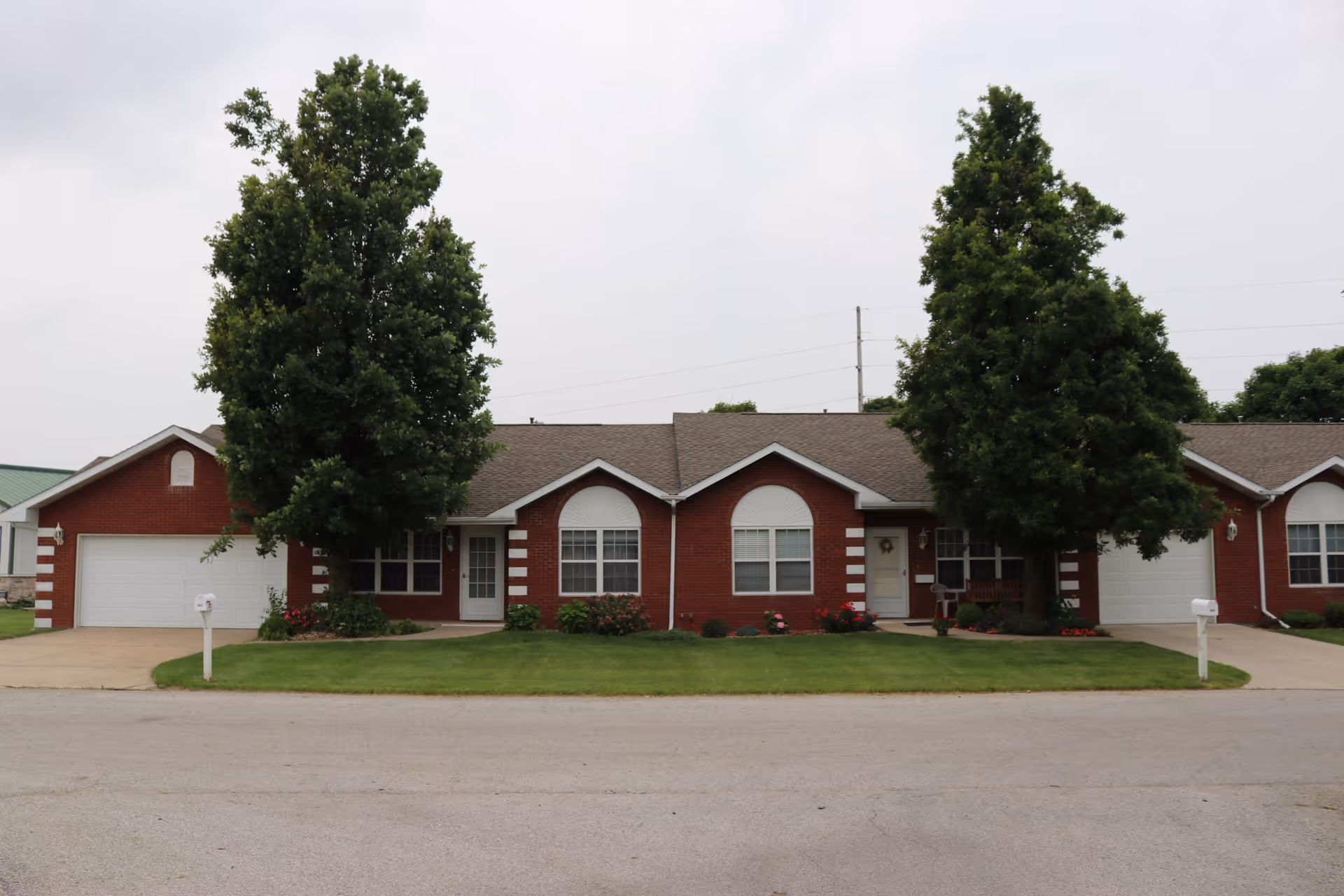 Front view of a single-story red brick senior living building with garages, two large trees, and a manicured lawn.