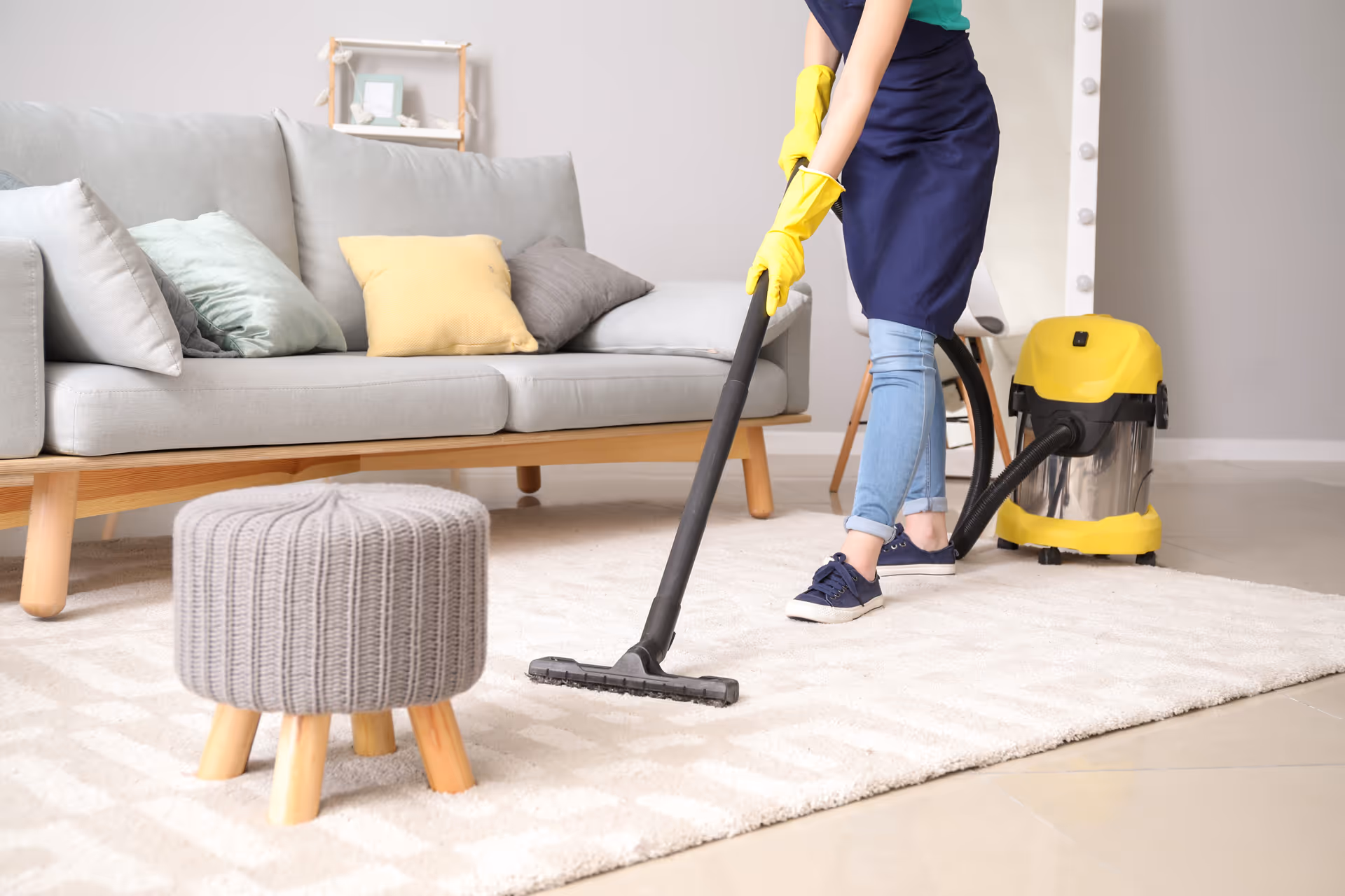 Person wearing gloves vacuums a light living room with sofa, rug, and ottoman.