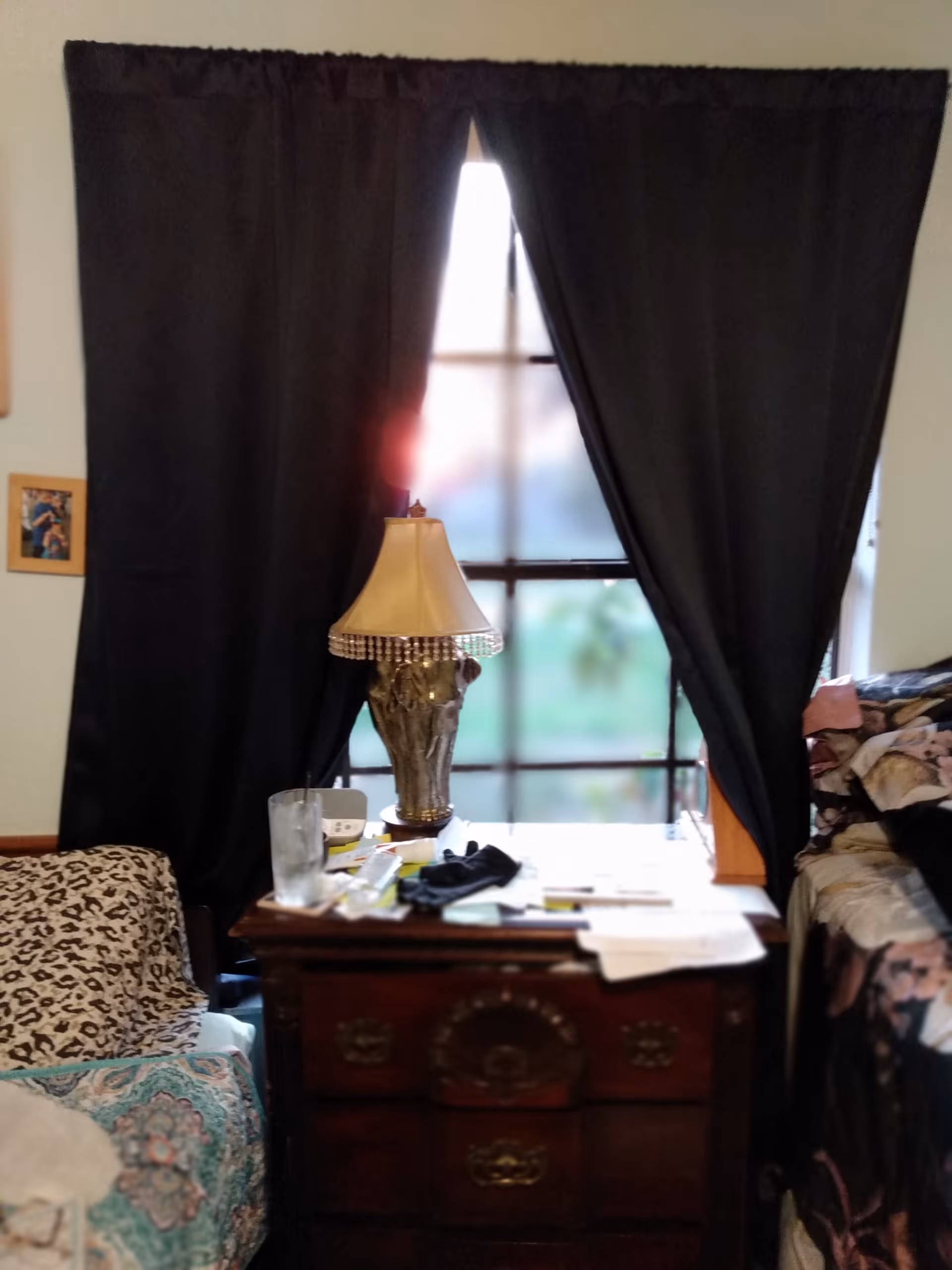 Dimly lit bedroom with dark curtains, a wooden dresser, leopard-print chair, and a bed.
