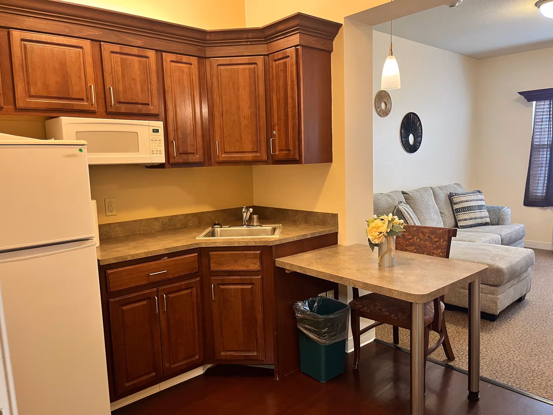 A small kitchen area with wooden cabinets, a white refrigerator, a microwave, and a sink. Adjacent to the kitchen is a small dining table with a floral centerpiece and a chair. In the background, there is a living room area with a light gray sectional sofa, decorative wall hangings, and a window with dark curtains.