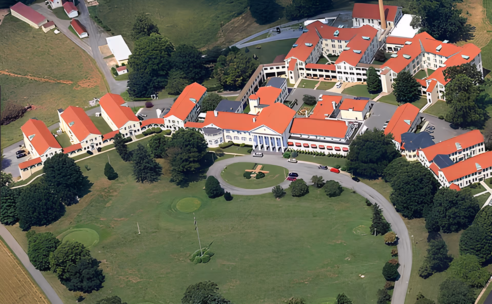 Aerial view of a large senior living facility with multiple connected buildings featuring red roofs, surrounded by green lawns, trees, and a circular driveway with parked cars.