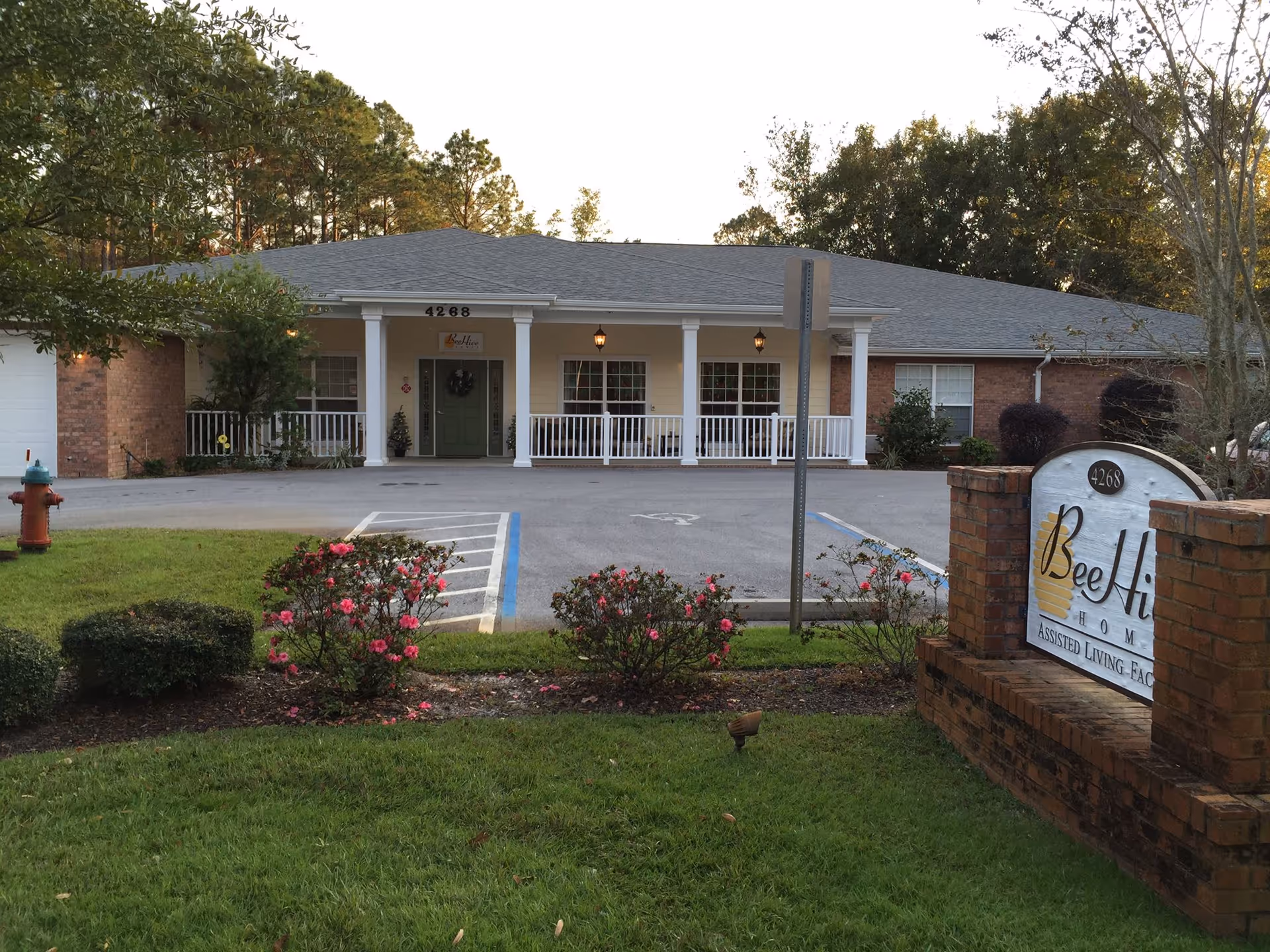 Exterior view of BeeHive Homes Assisted Living facility showing a single-story building with a covered porch supported by white columns. There is a parking area with handicap spaces in front, surrounded by green grass, bushes, and flowering plants. A brick sign with the facility name and address is visible on the right side of the image.
