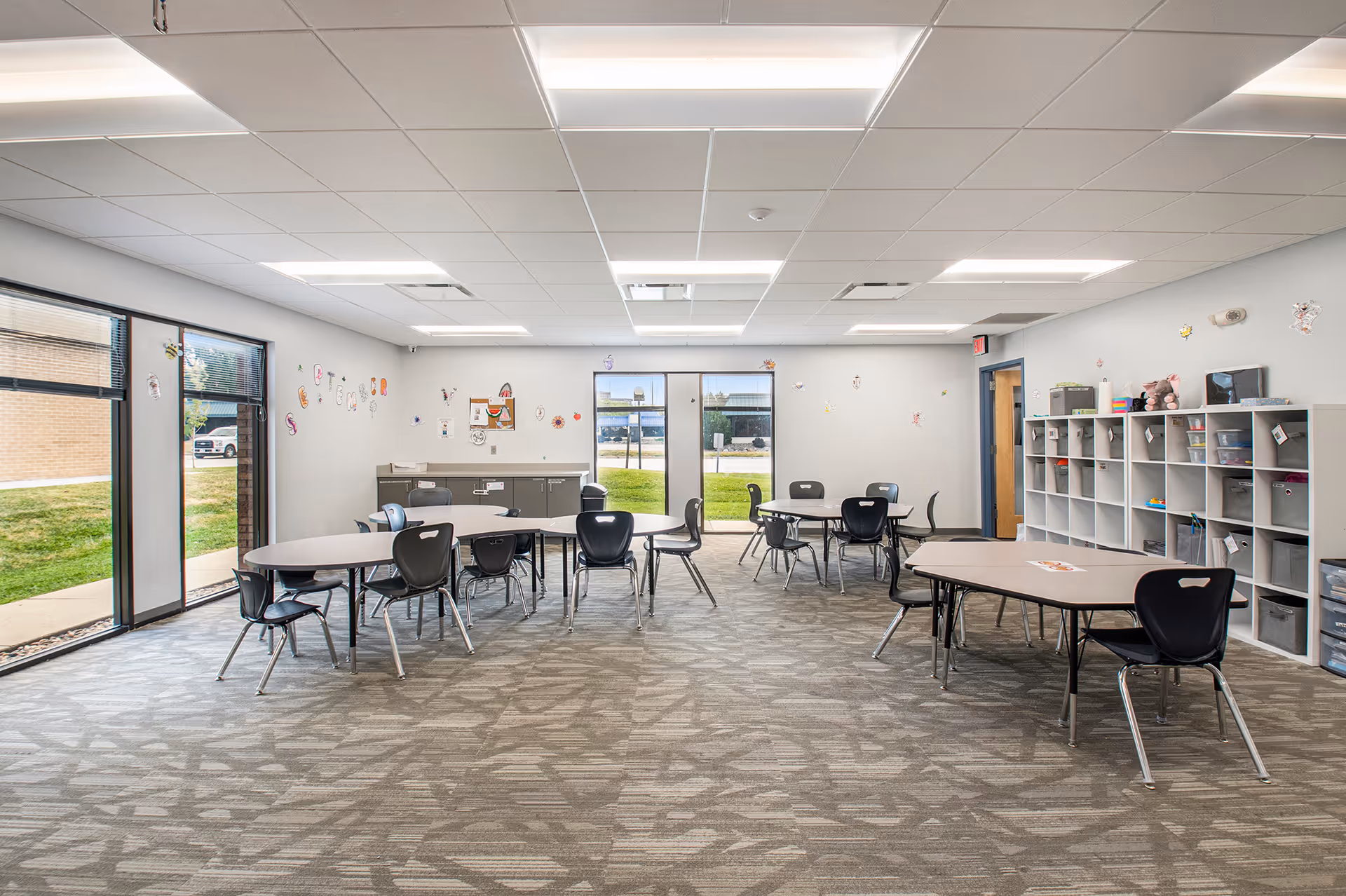Bright indoor activity room with round tables, chairs, and cubbies along a wall.