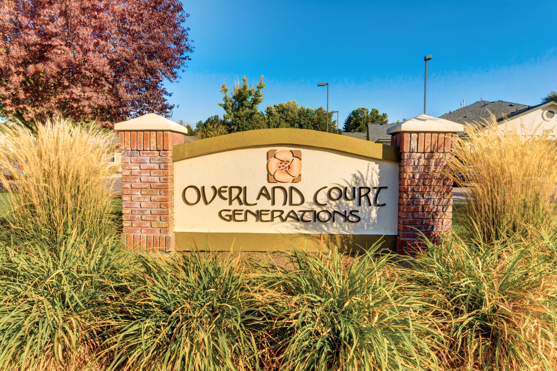 Exterior entrance sign for Overland Court Generations, framed by brick pillars and tall grasses.