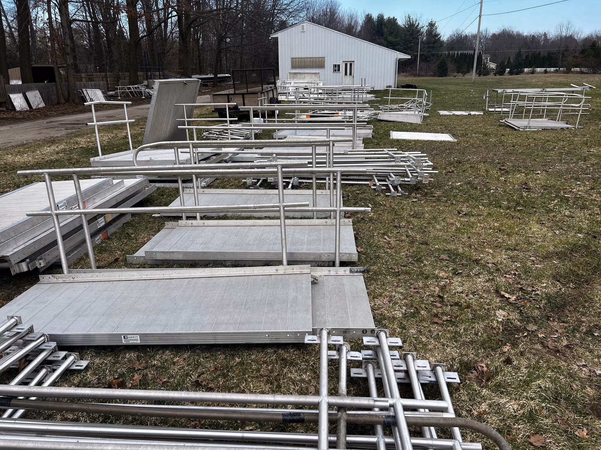 Metal portable ramps and rails are stacked outdoors on a grassy area near a white building.