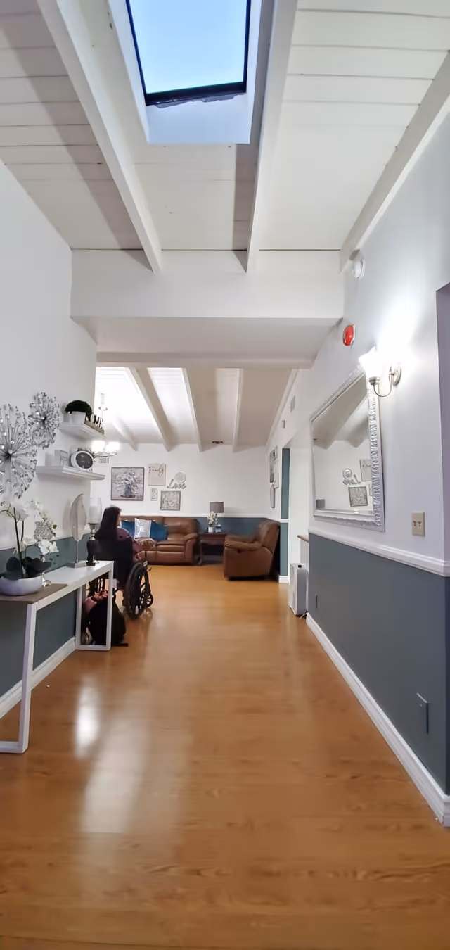 A bright hallway with wooden flooring and white walls with a blue lower half. There is a skylight on the ceiling allowing natural light in. On the left side, a woman in a wheelchair is positioned near a white console table decorated with plants and ornaments. At the end of the hallway, there is a cozy seating area with brown leather sofas, a side table, and wall art including framed pictures and decorative words.