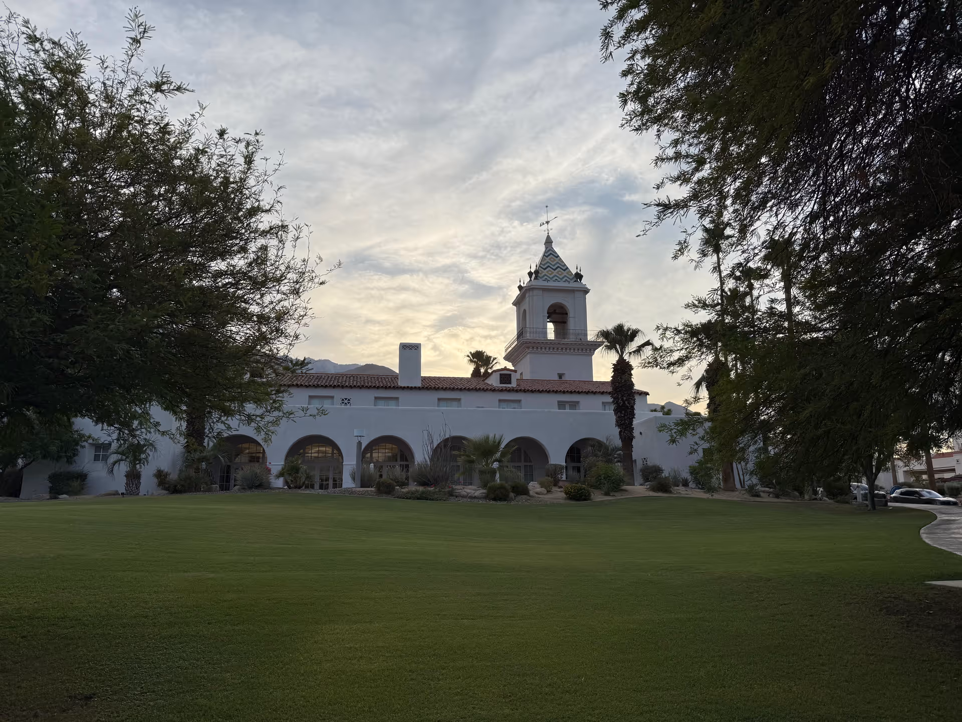 White Mediterranean-style building with a tower, arches, palm trees, and a grassy front lawn.