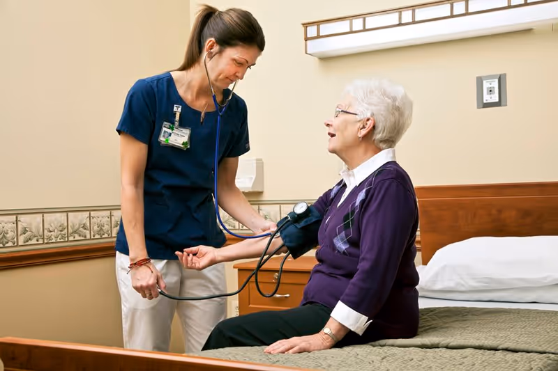 A healthcare worker in navy scrubs is measuring the blood pressure of an elderly woman with white hair who is sitting on a bed in a room. The elderly woman is wearing glasses and a purple sweater with an argyle pattern. The room has beige walls and a wooden headboard with a white pillow and green blanket on the bed.