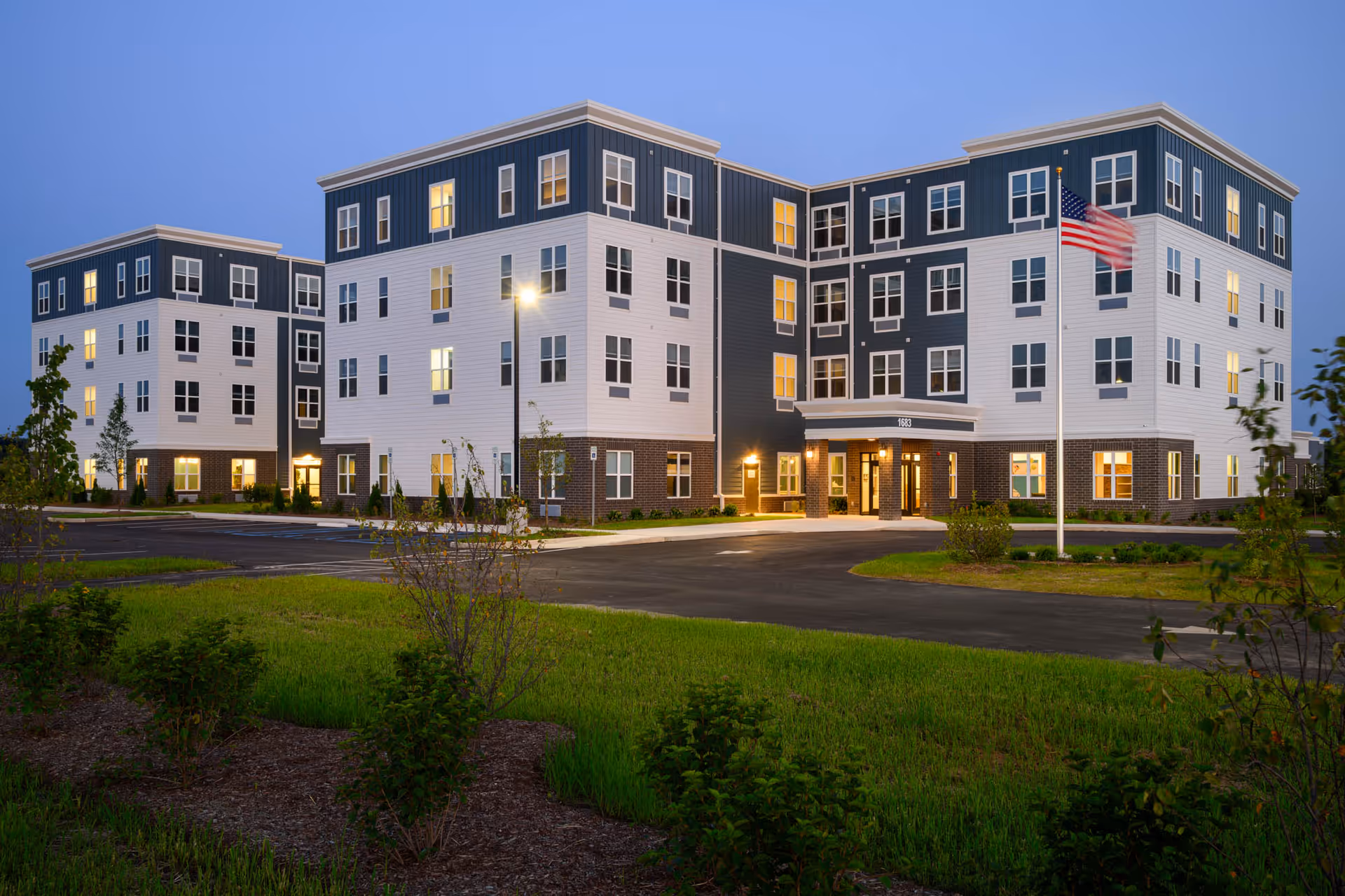 Modern multi-story senior living facility exterior with flag, lit entrances, and landscaping at dusk.