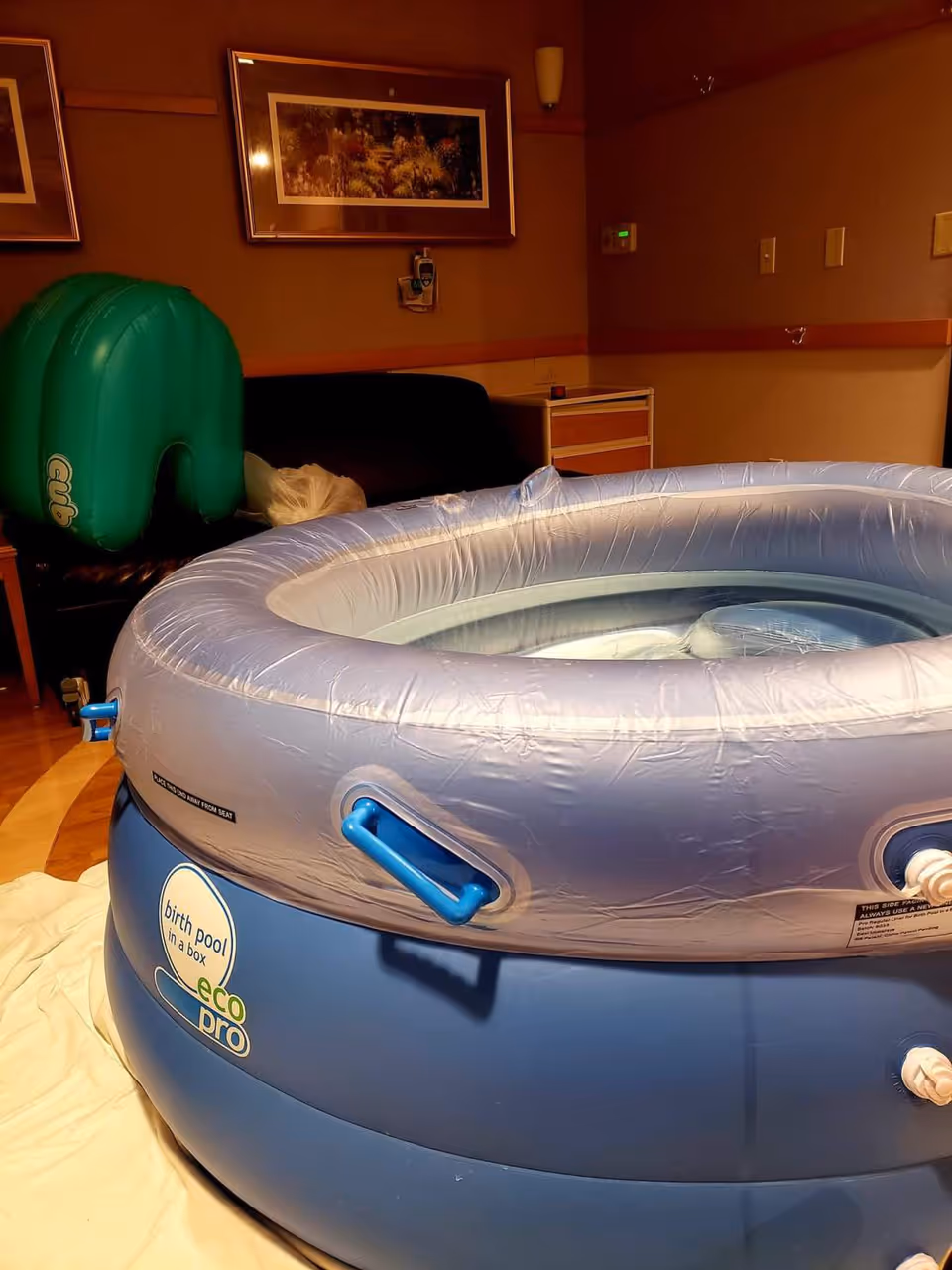 Indoor inflatable birth pool in a dim room with equipment and framed art.